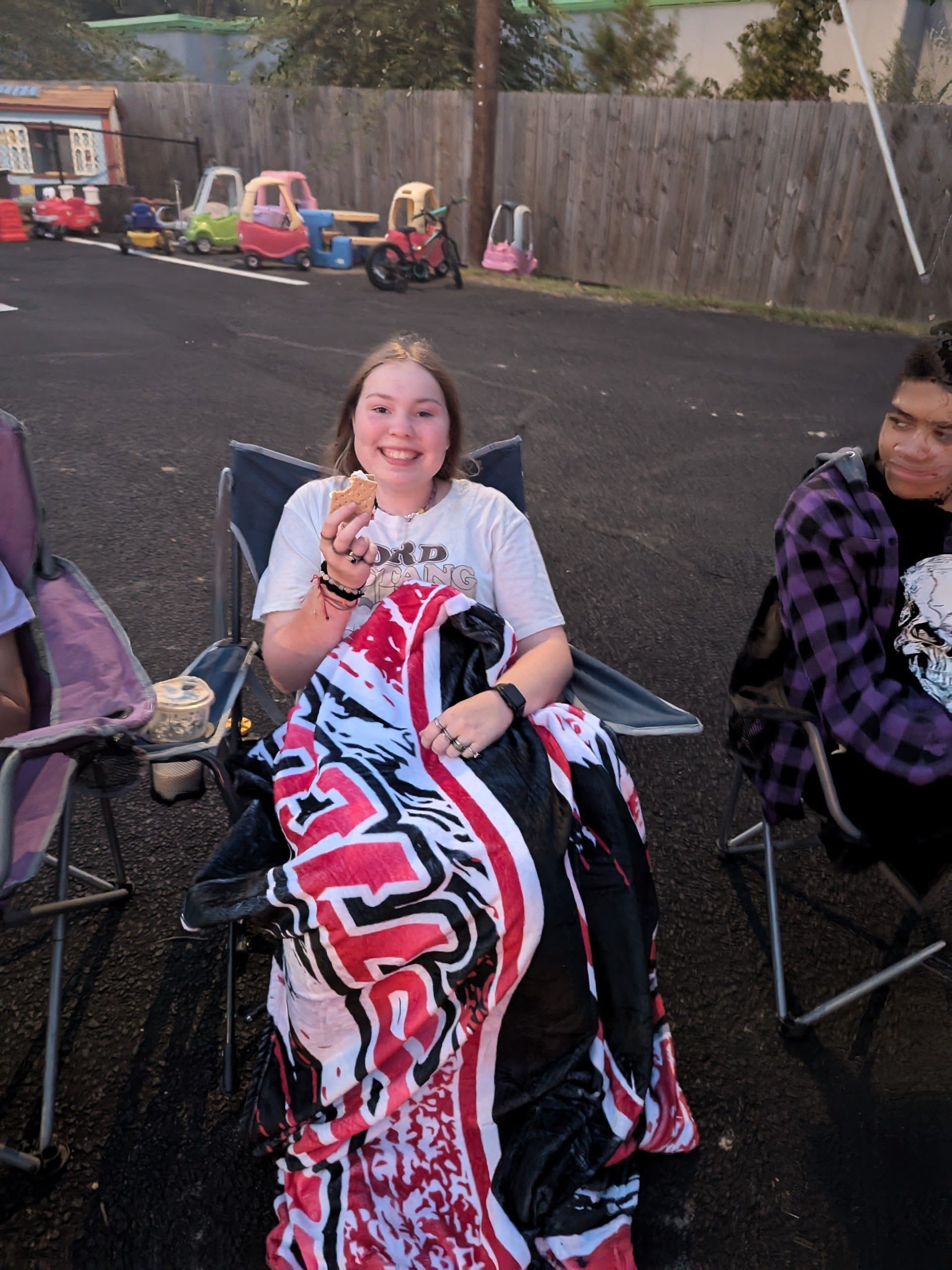 A smiling person eats a snack while wrapped in a blanket, sitting in a chair. Another person is seated nearby.