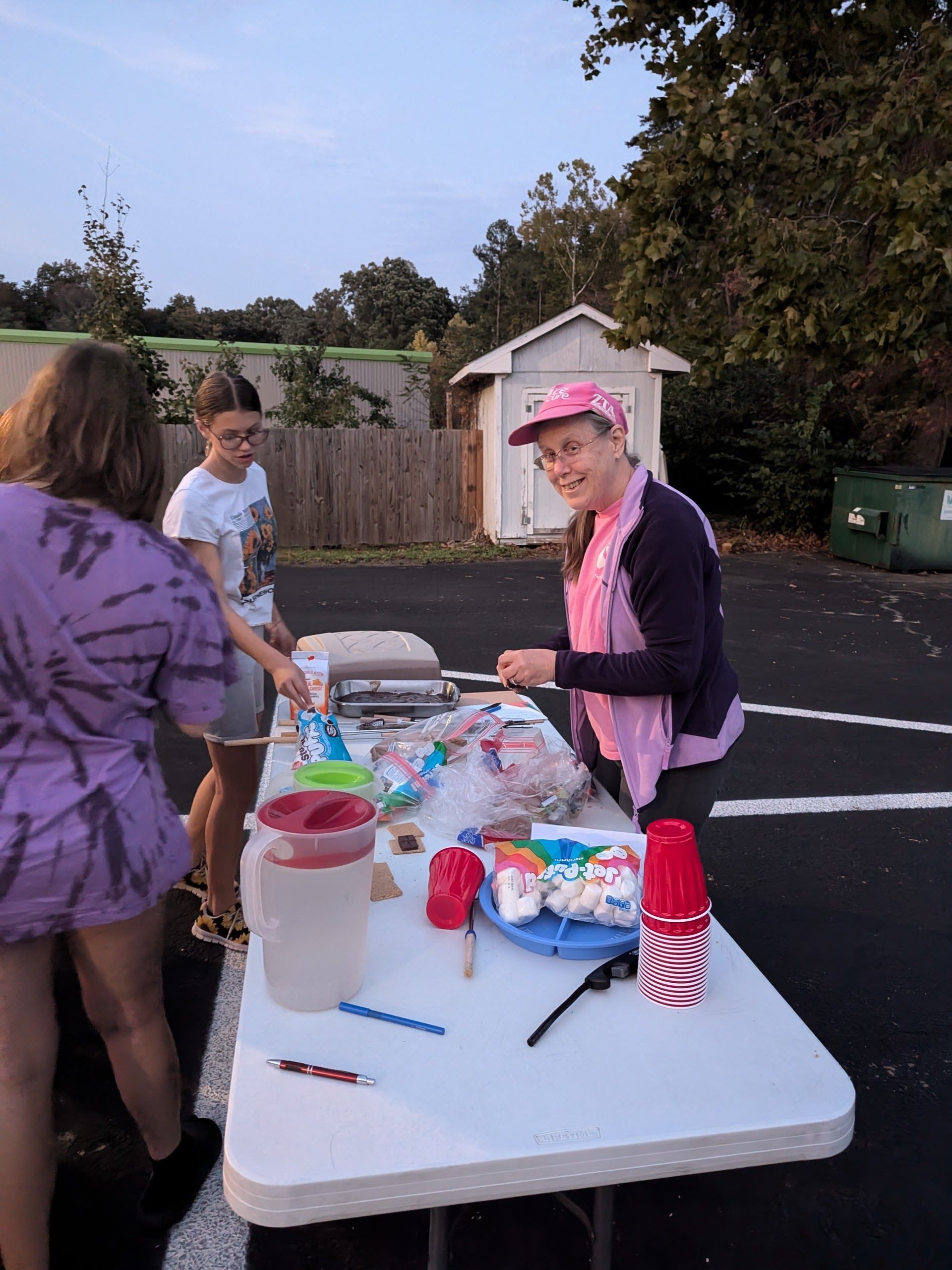 People at a table with drinks and snacks, outside. Woman in pink smiles. Two girls assist.