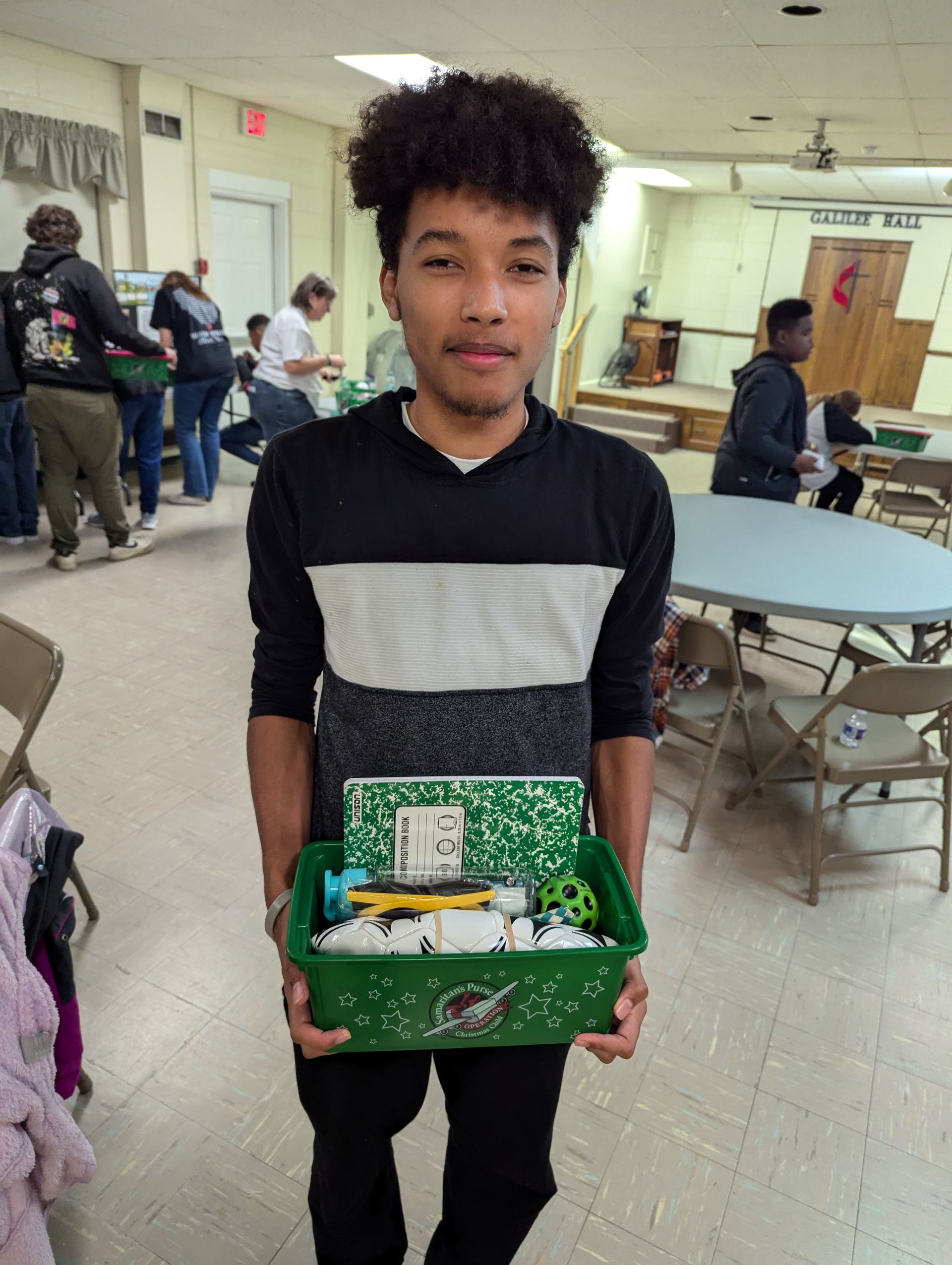 Young man holds a green bin filled with school supplies in a community center.