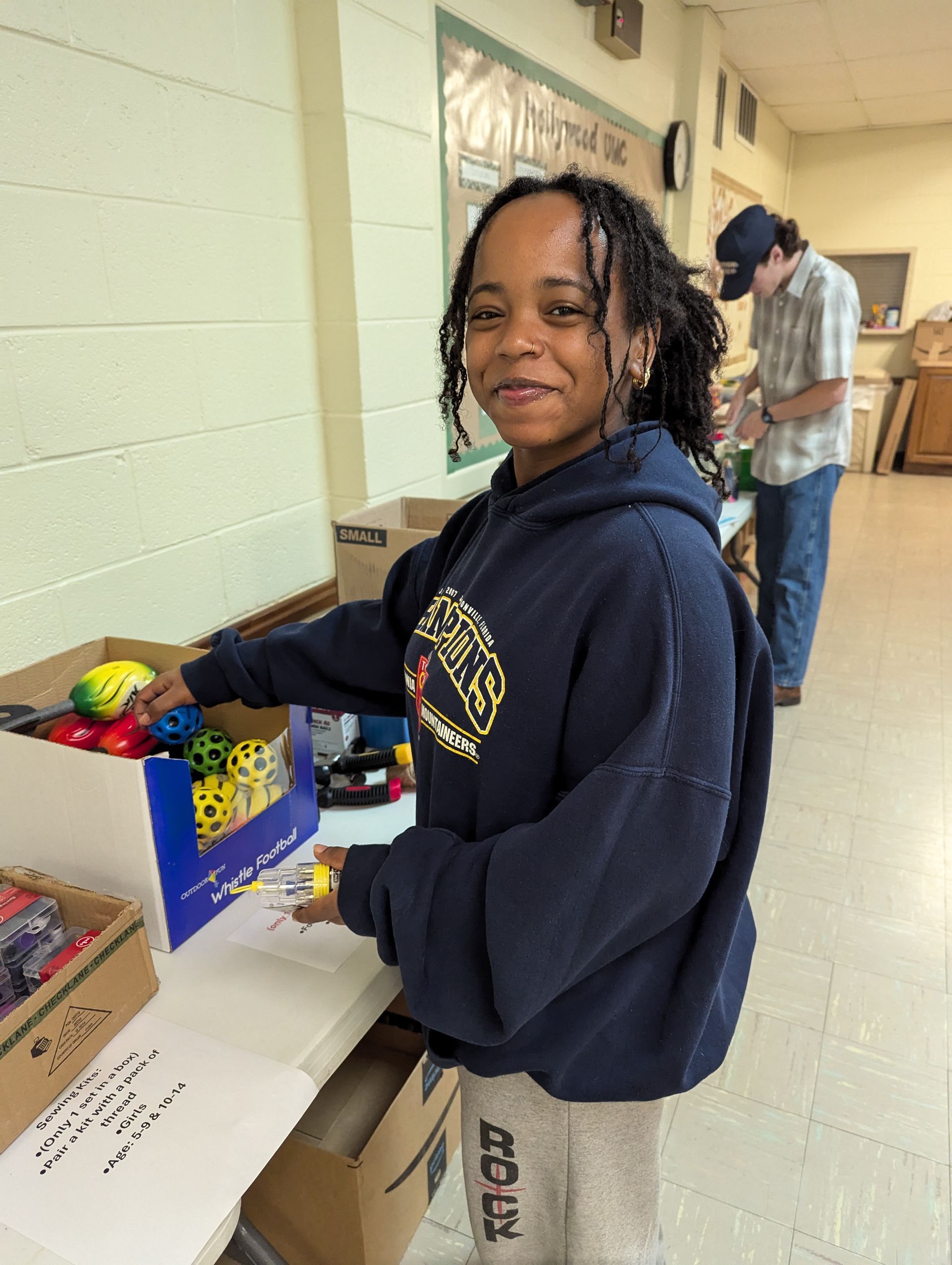Young woman sorting toys in a room. She smiles, wearing a blue hoodie. Another person stands in the background.