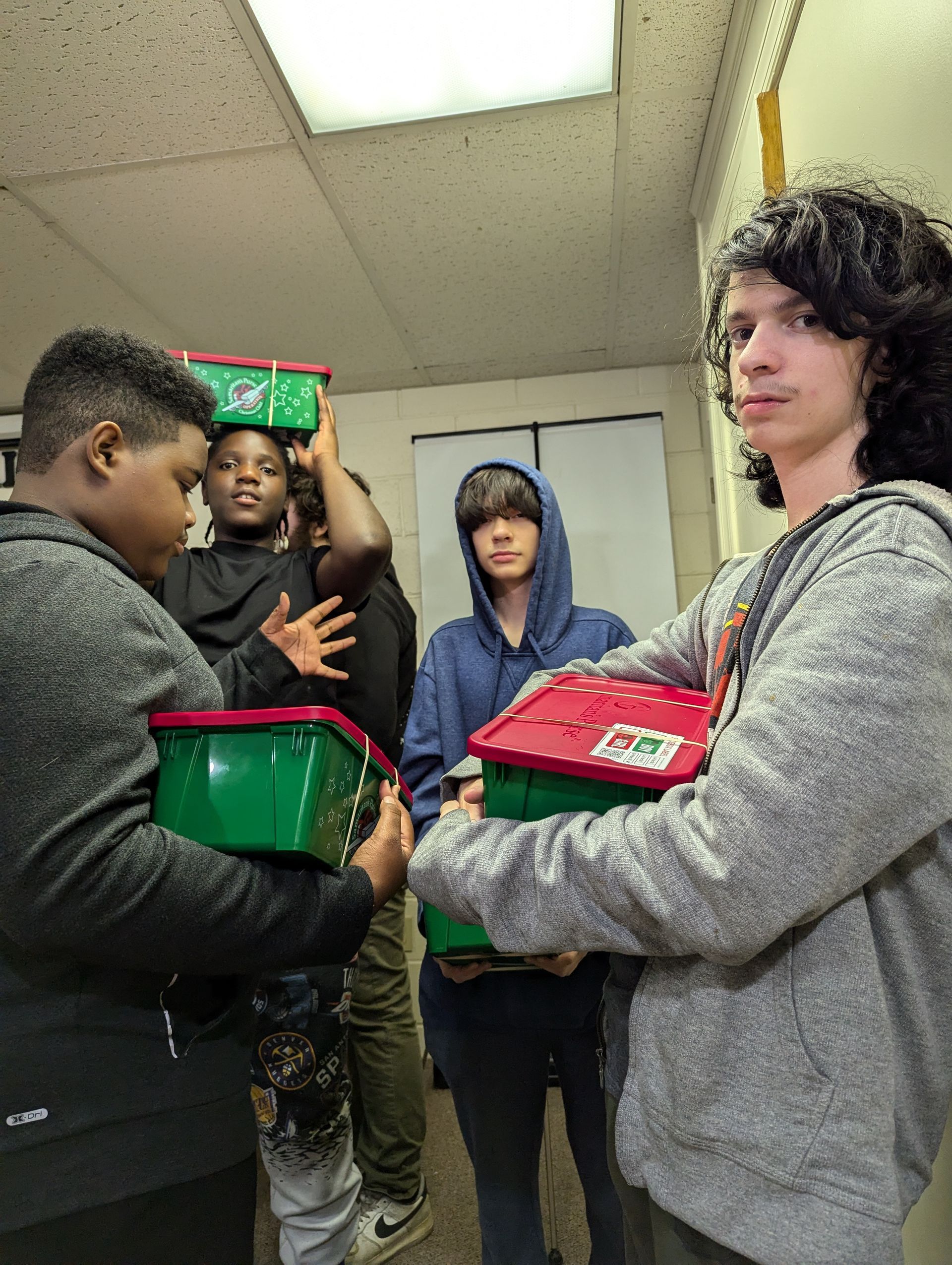 Five people, holding and balancing holiday boxes, in an indoor setting. Some wear hoodies; one has curly hair.