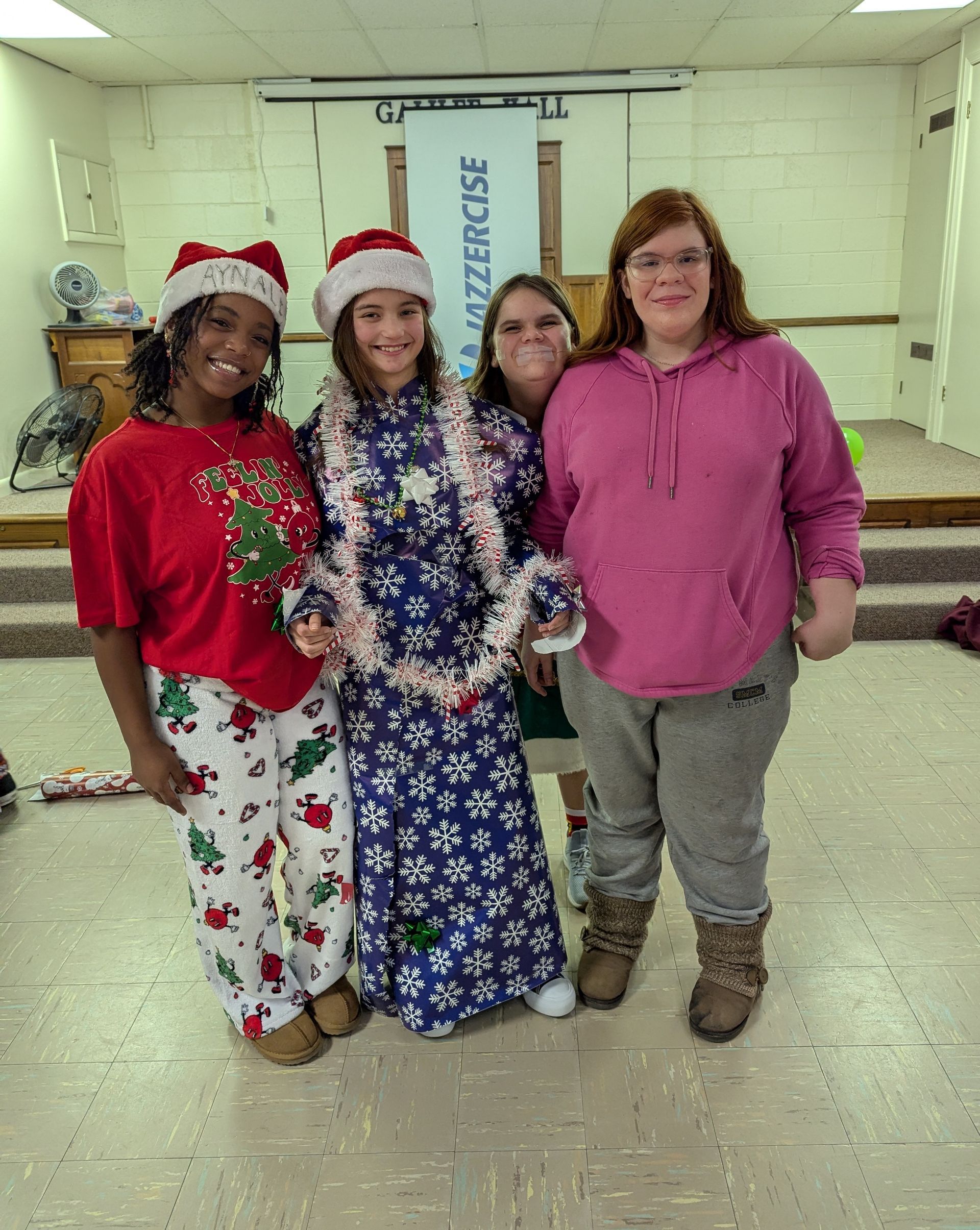 Four people in festive attire pose indoors, two in Santa hats.