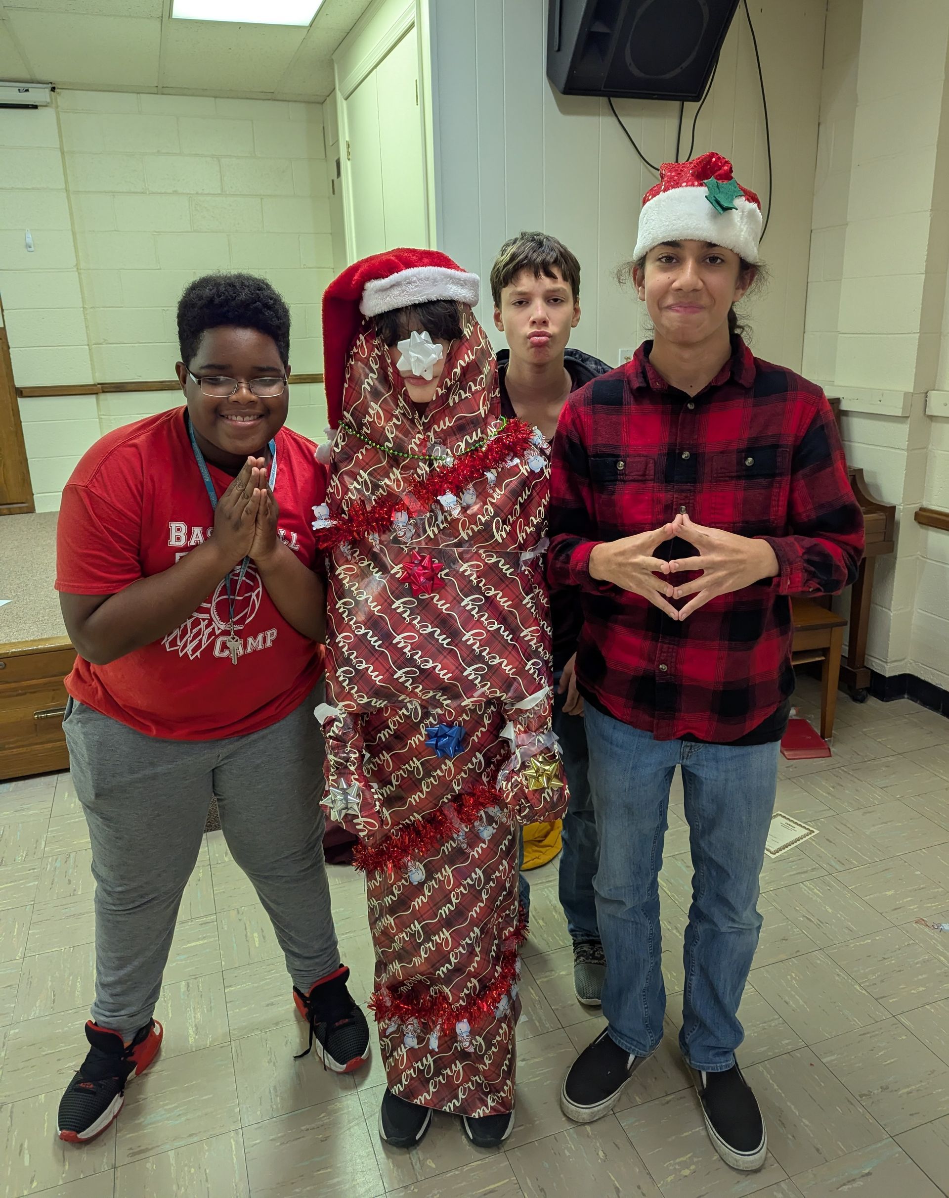 Four people, one wrapped in holiday paper, pose. Two in red, one in a Santa hat, other two smiling in a room.