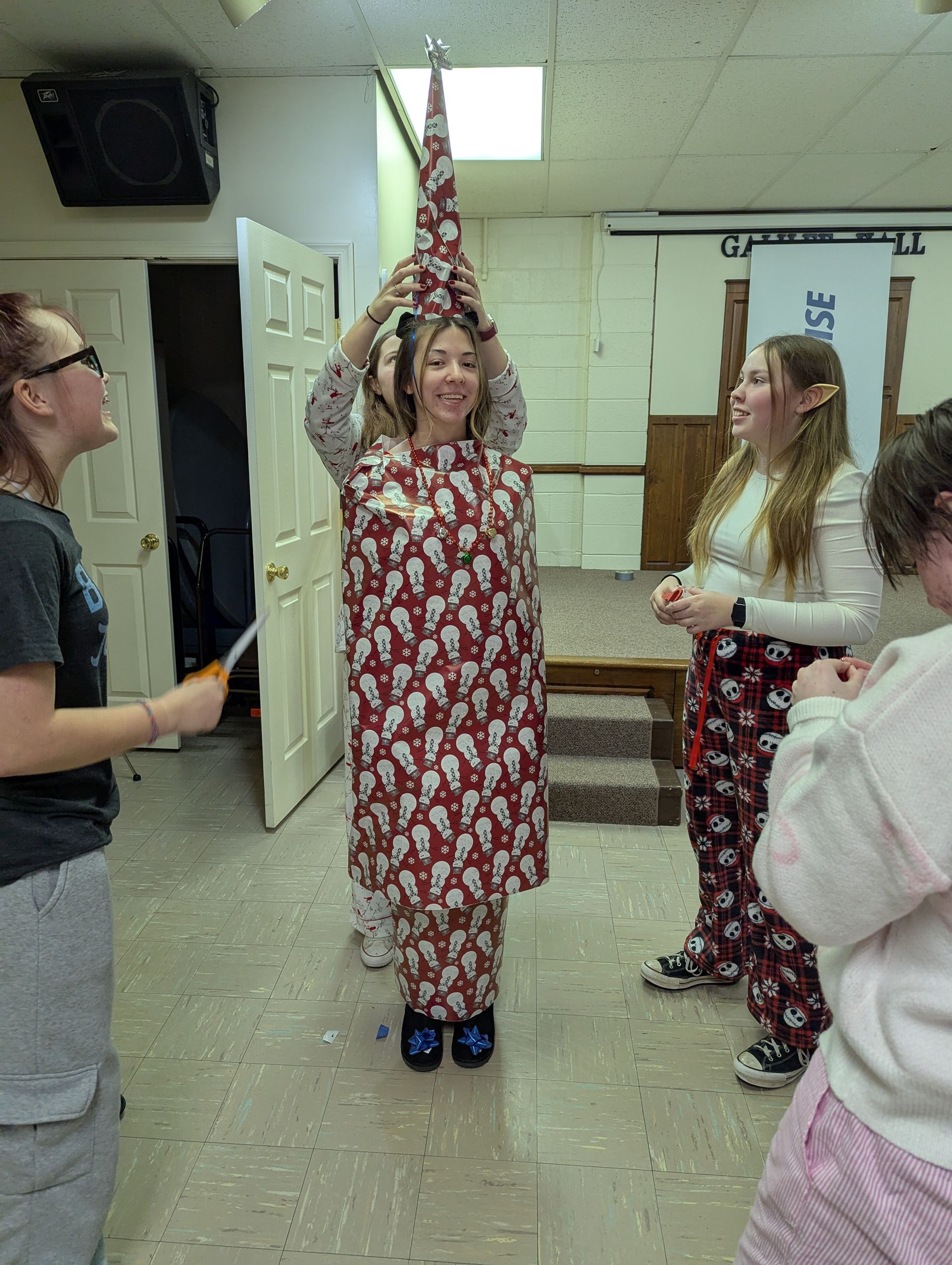 Four people wrap a person in patterned paper to look like a Christmas tree, indoors.