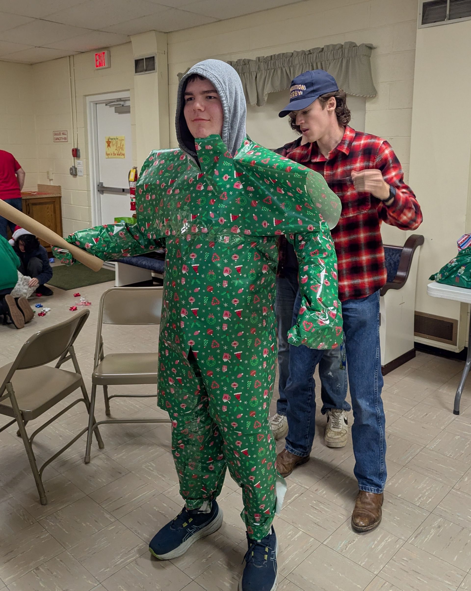 Person in green Christmas onesie being adjusted by a person in a plaid shirt. Inside a room.