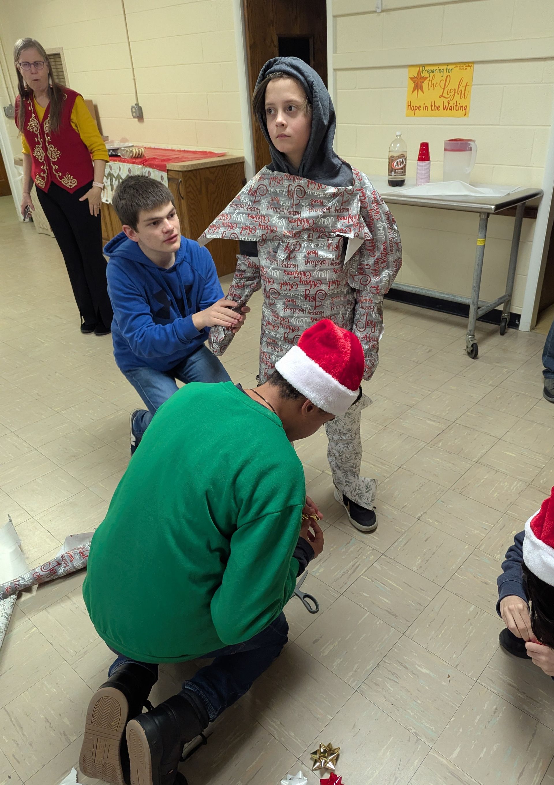 Children wrapping another child in wrapping paper, one kneeling. Others watching, wearing Santa hats, in a room.