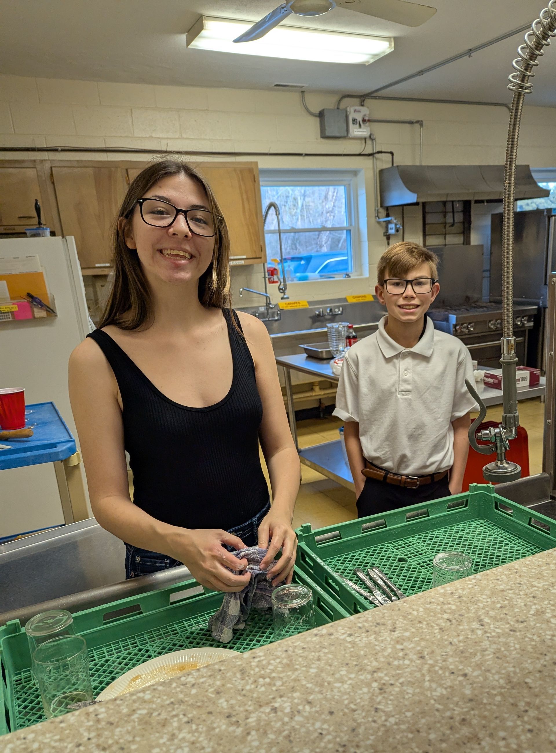 Two young people smiling in a kitchen, near dishwashing area. One washes dishes, other stands by.