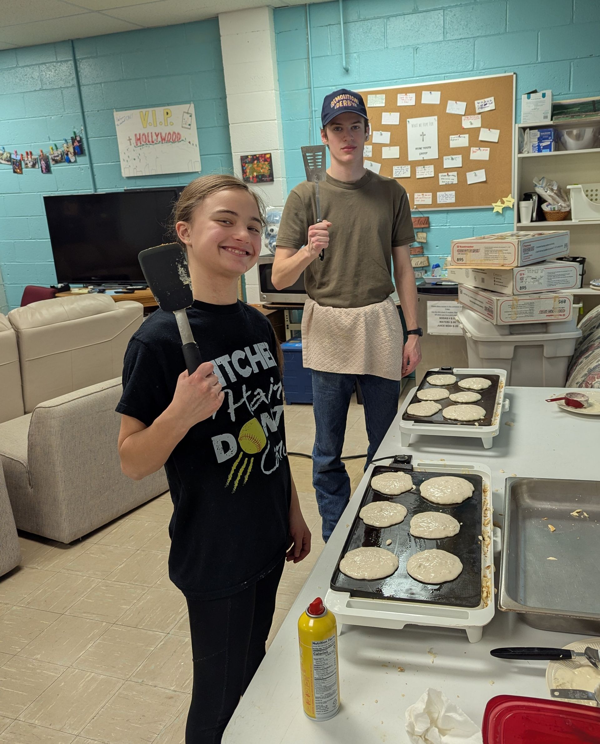 Two people flipping pancakes on griddles. Teen girl smiles, holding a spatula. Teen boy holds a fork.