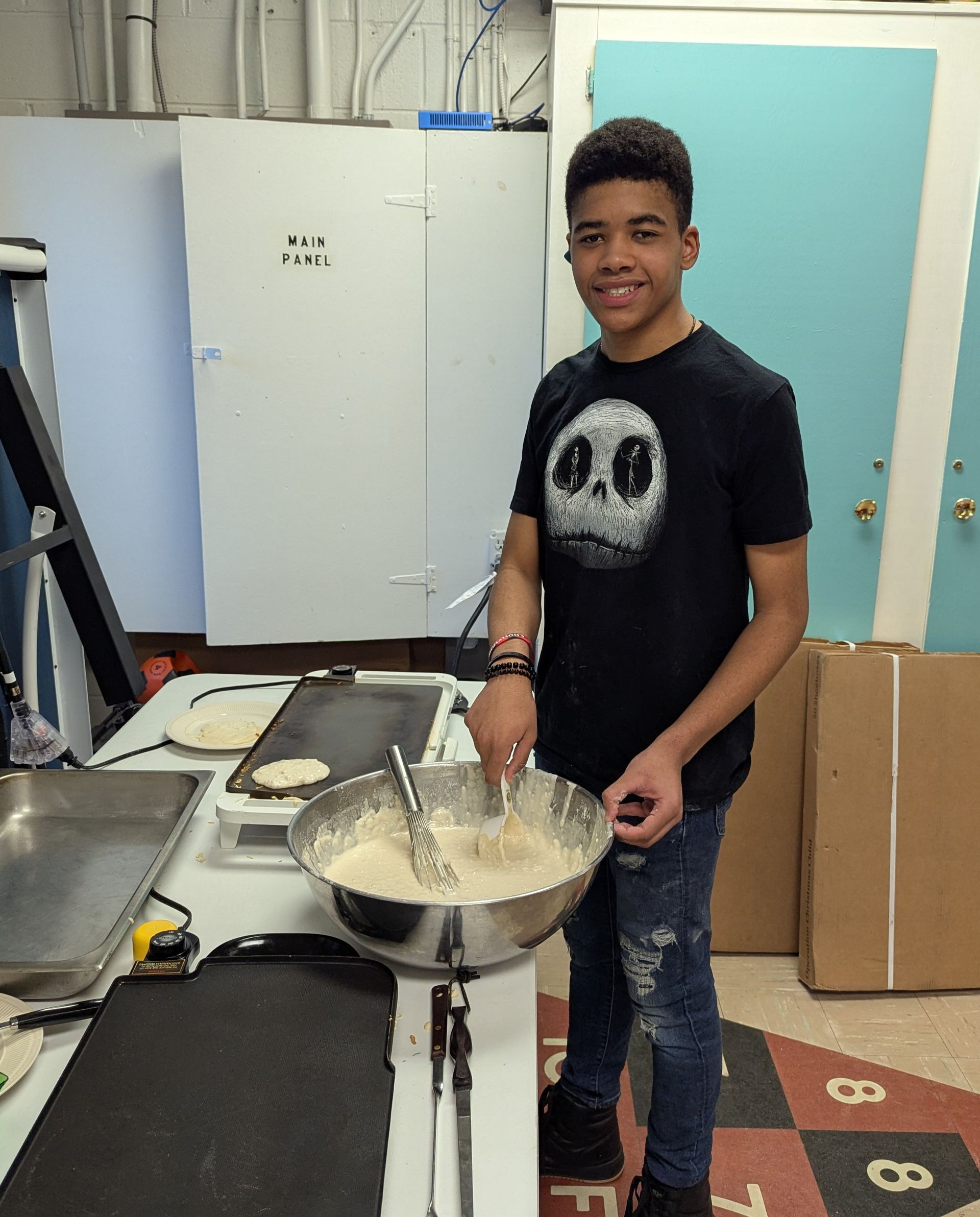 Teenager mixing batter, smiling, in kitchen; griddle and cabinets in background.