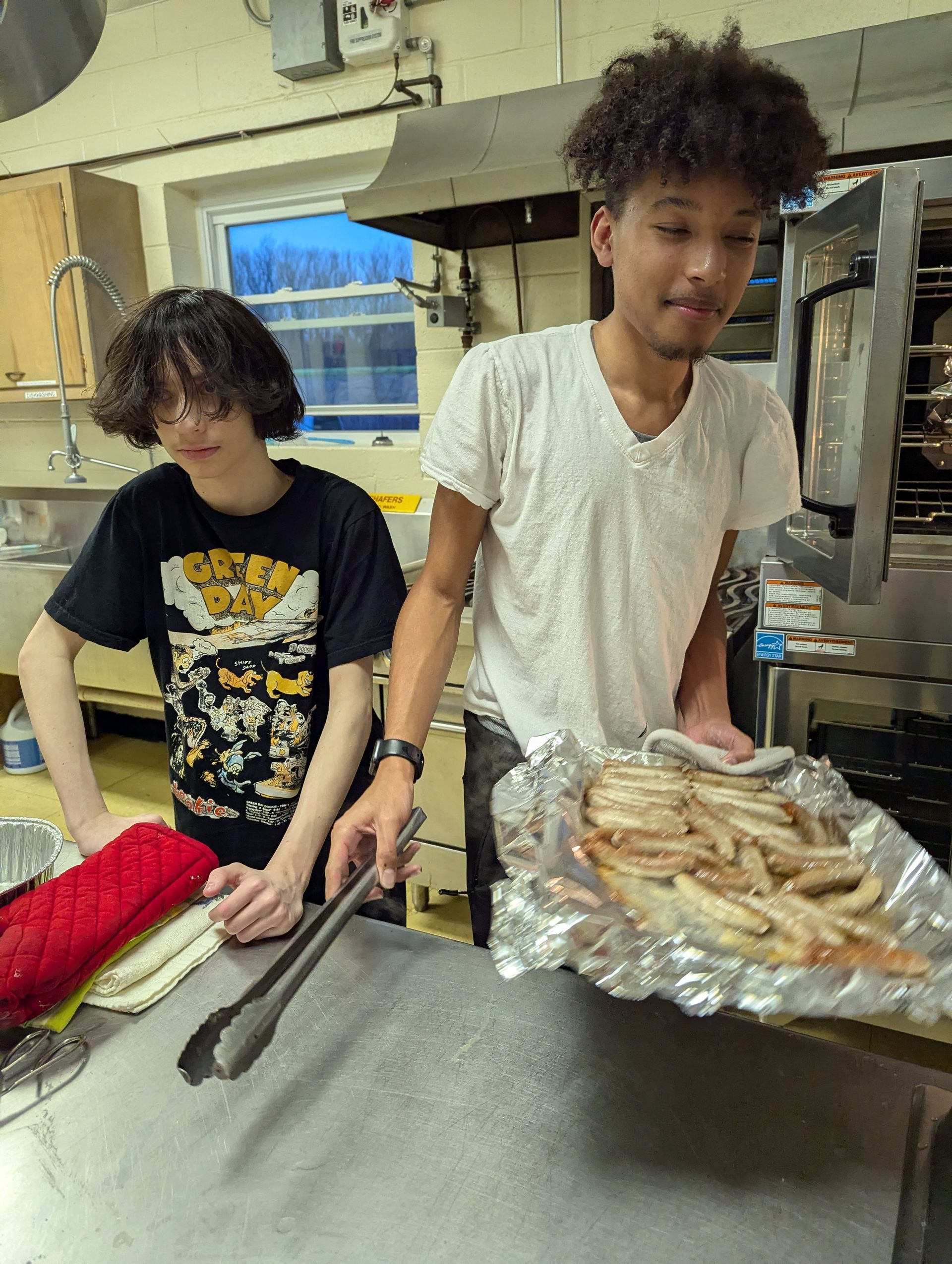 Two young people in a kitchen; one holds fries in foil, the other uses tongs.
