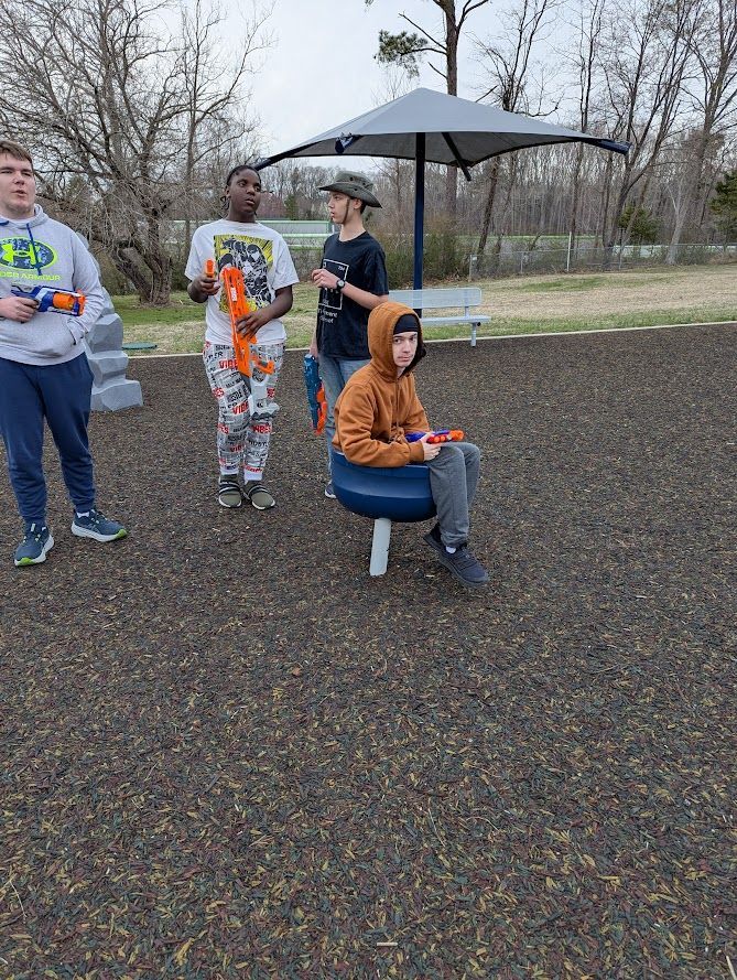 Four people holding Nerf guns on a playground, one sitting on a spinner.