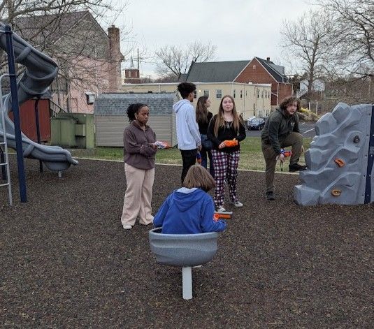 A group of teens at a playground. One is in a spinning seat while others stand nearby, some with hands out.
