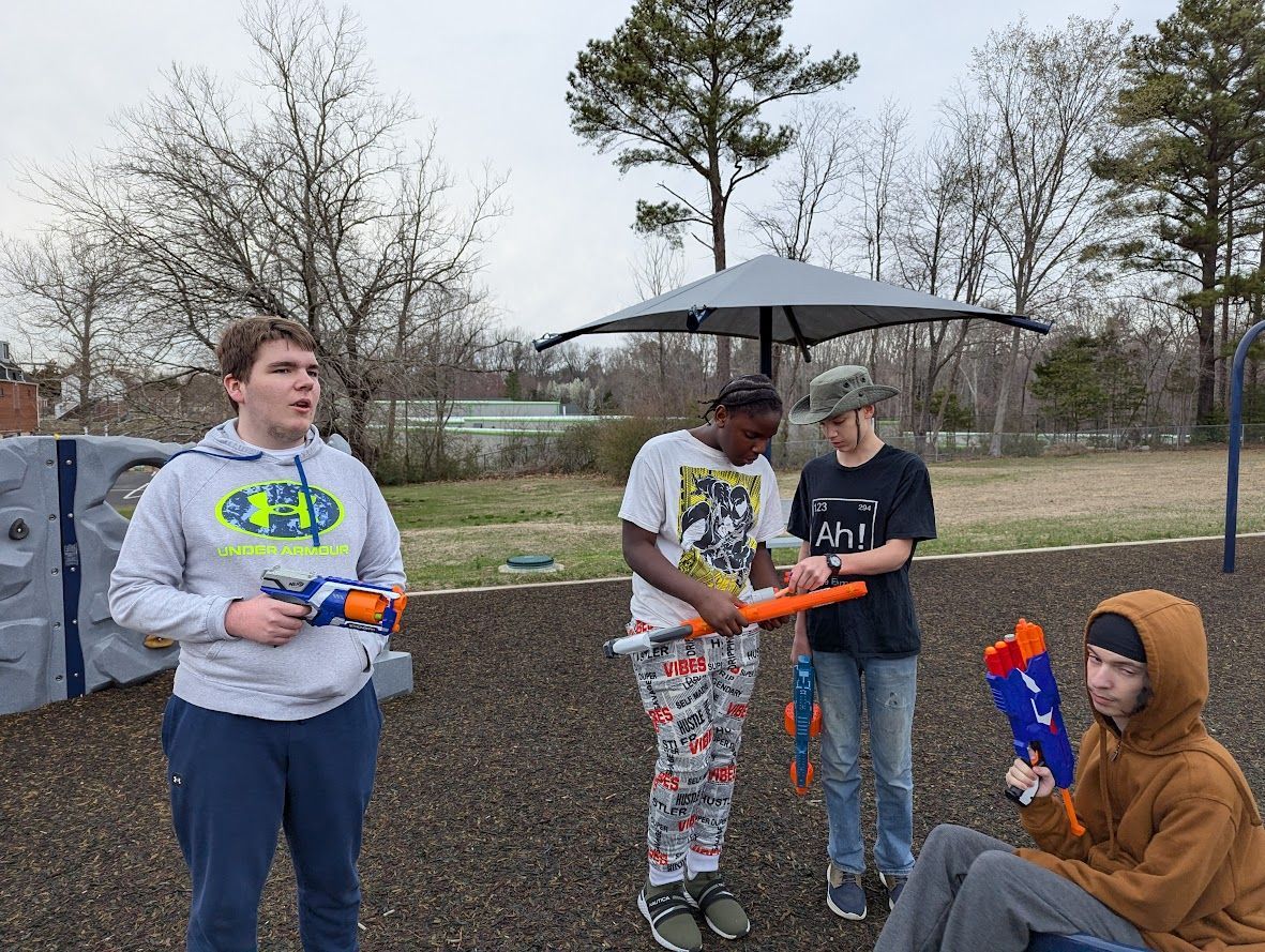Four people with Nerf guns outdoors, two standing, two seated, under a gray umbrella, brown ground.