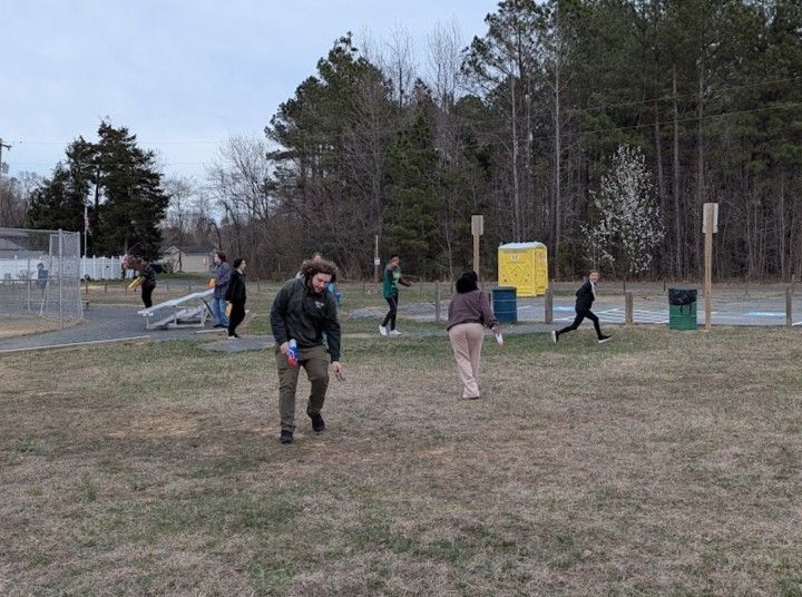 People playing outside; a man in a green jacket is in focus. There's a port-a-potty and trees in the background.