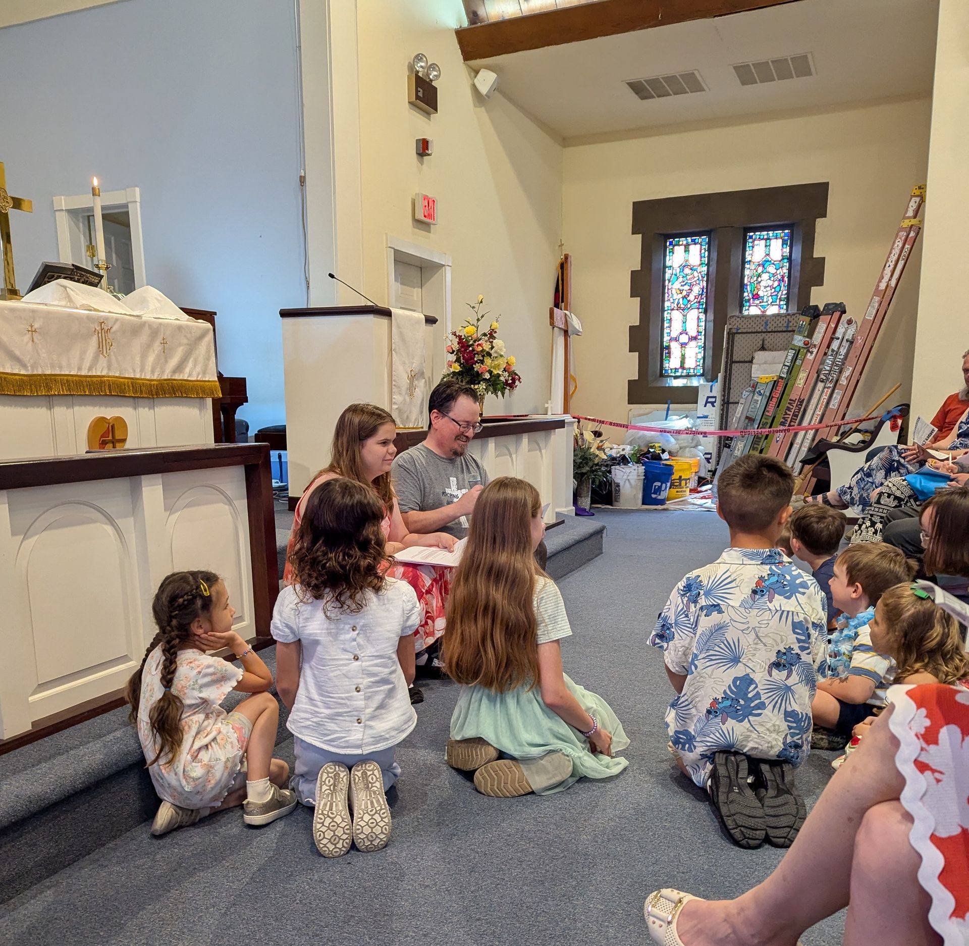 Children sit on the floor in a church, listening to an adult speak.