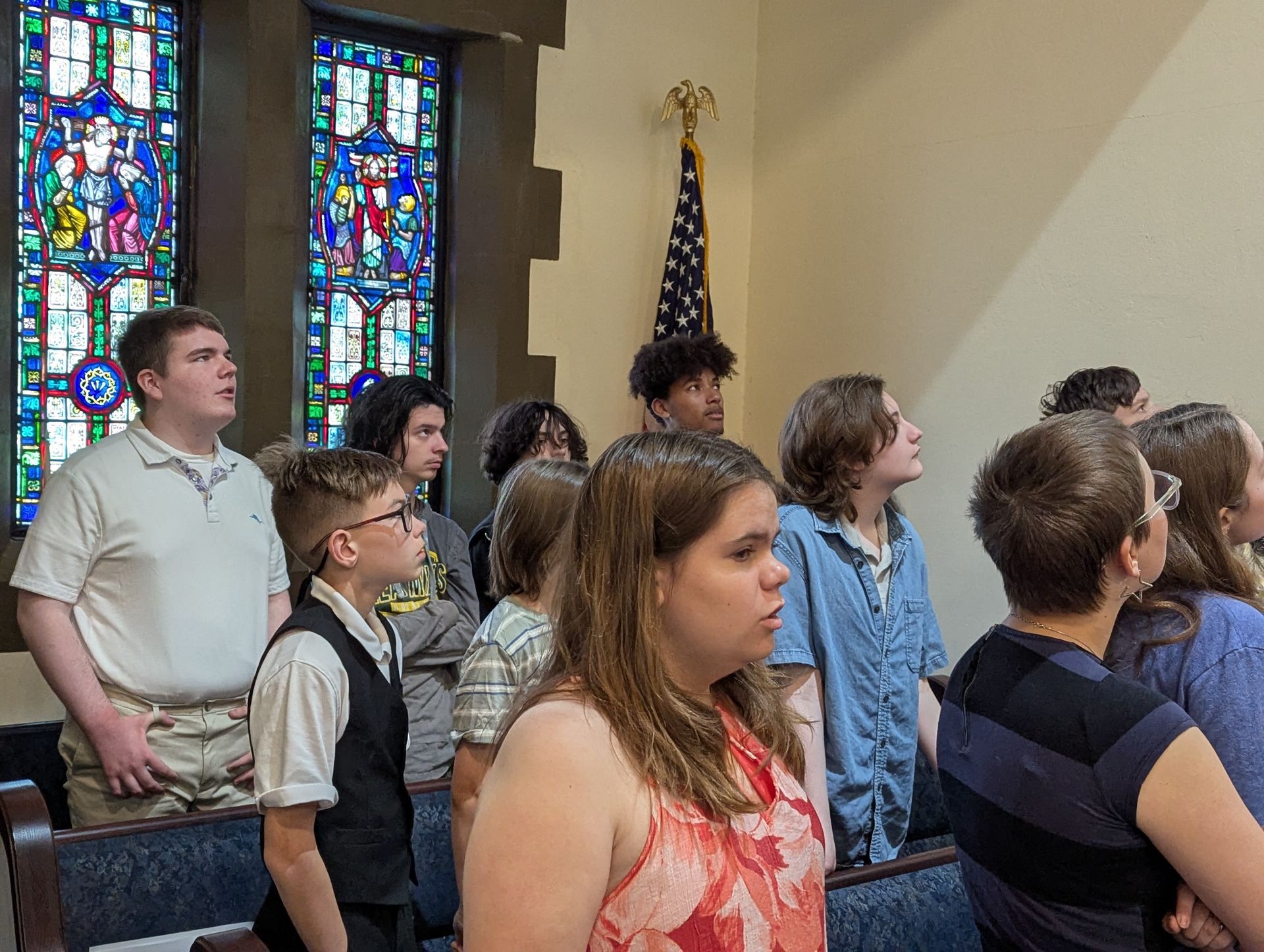 A group of diverse teens singing in a church. Stained glass windows and an American flag are visible.