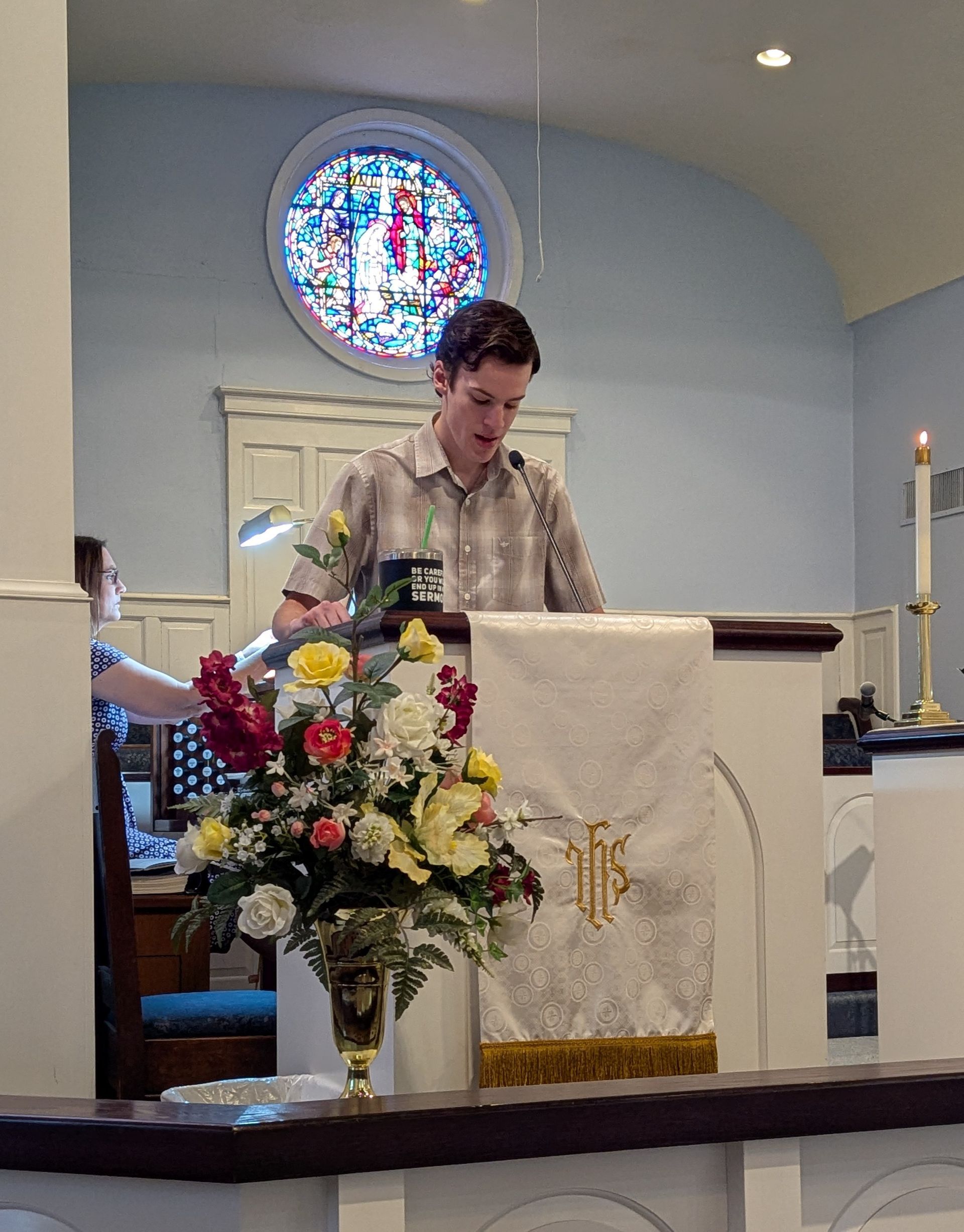 A youth in church giving a sermon, flowers on podium, stained glass in background.