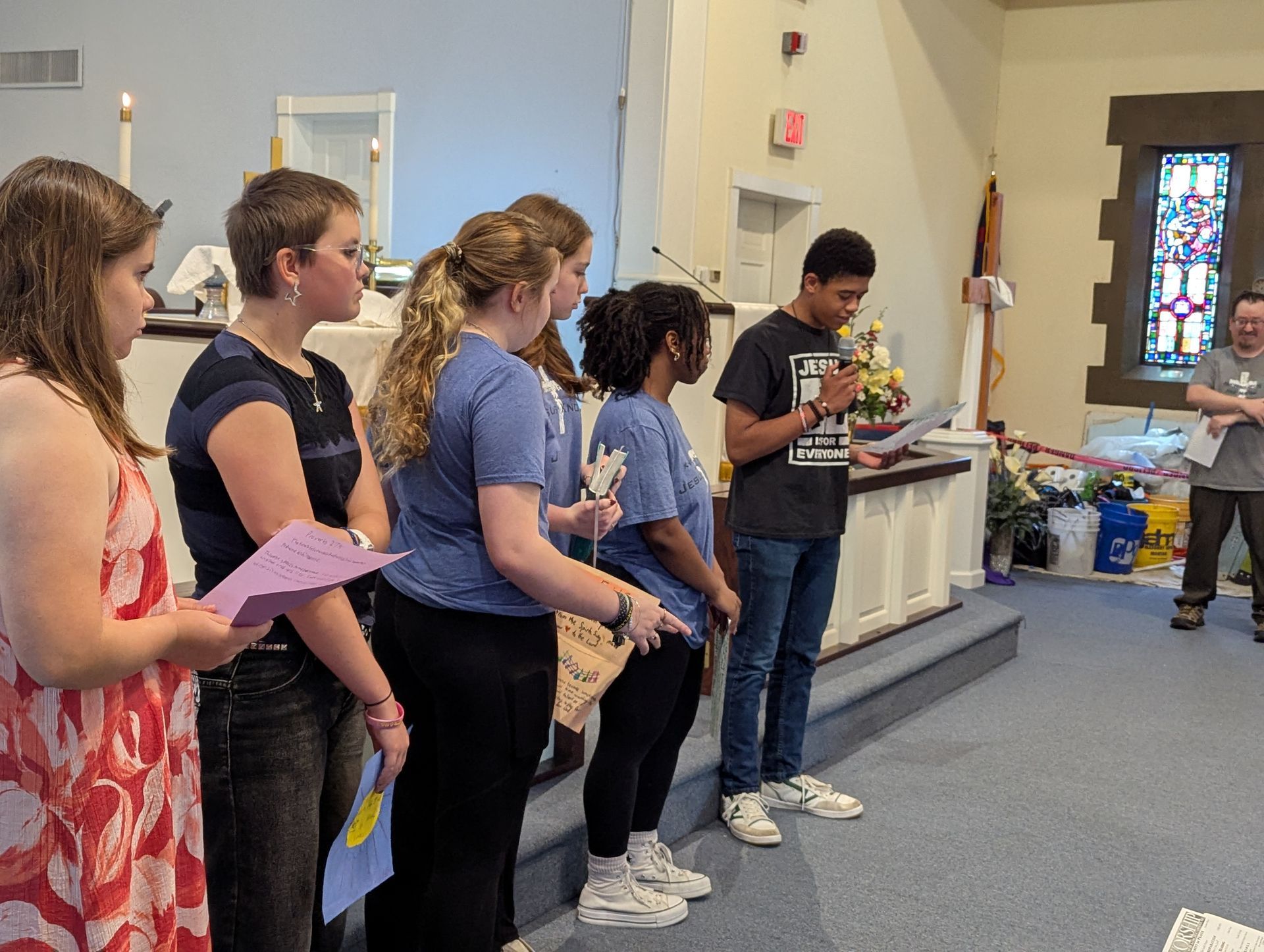 Group of teens speaking in a church. One boy holds a microphone, others hold papers and stand on steps.
