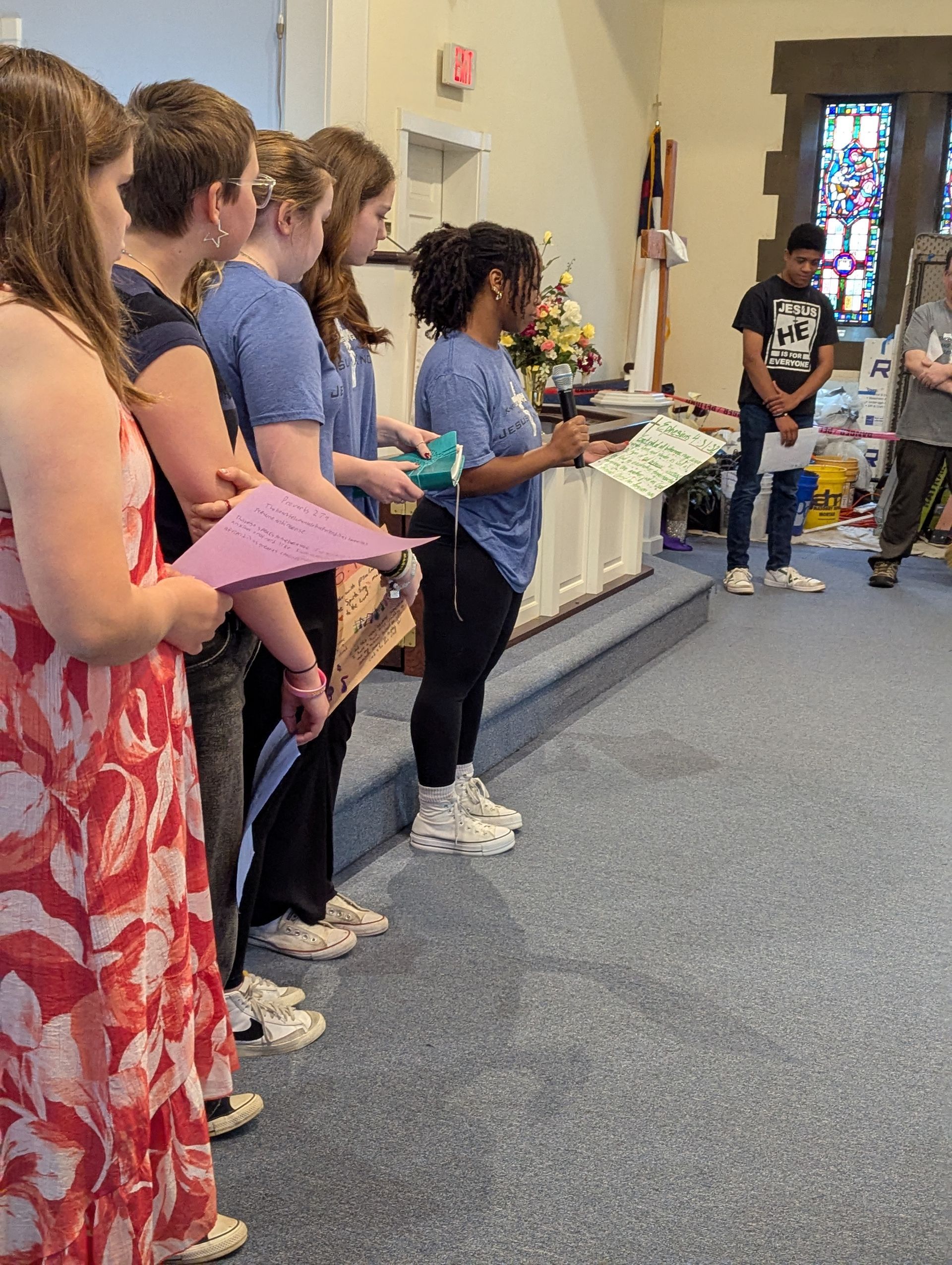 A group of young people in a church, reading from papers. Several wear blue shirts, and the room has stained glass.
