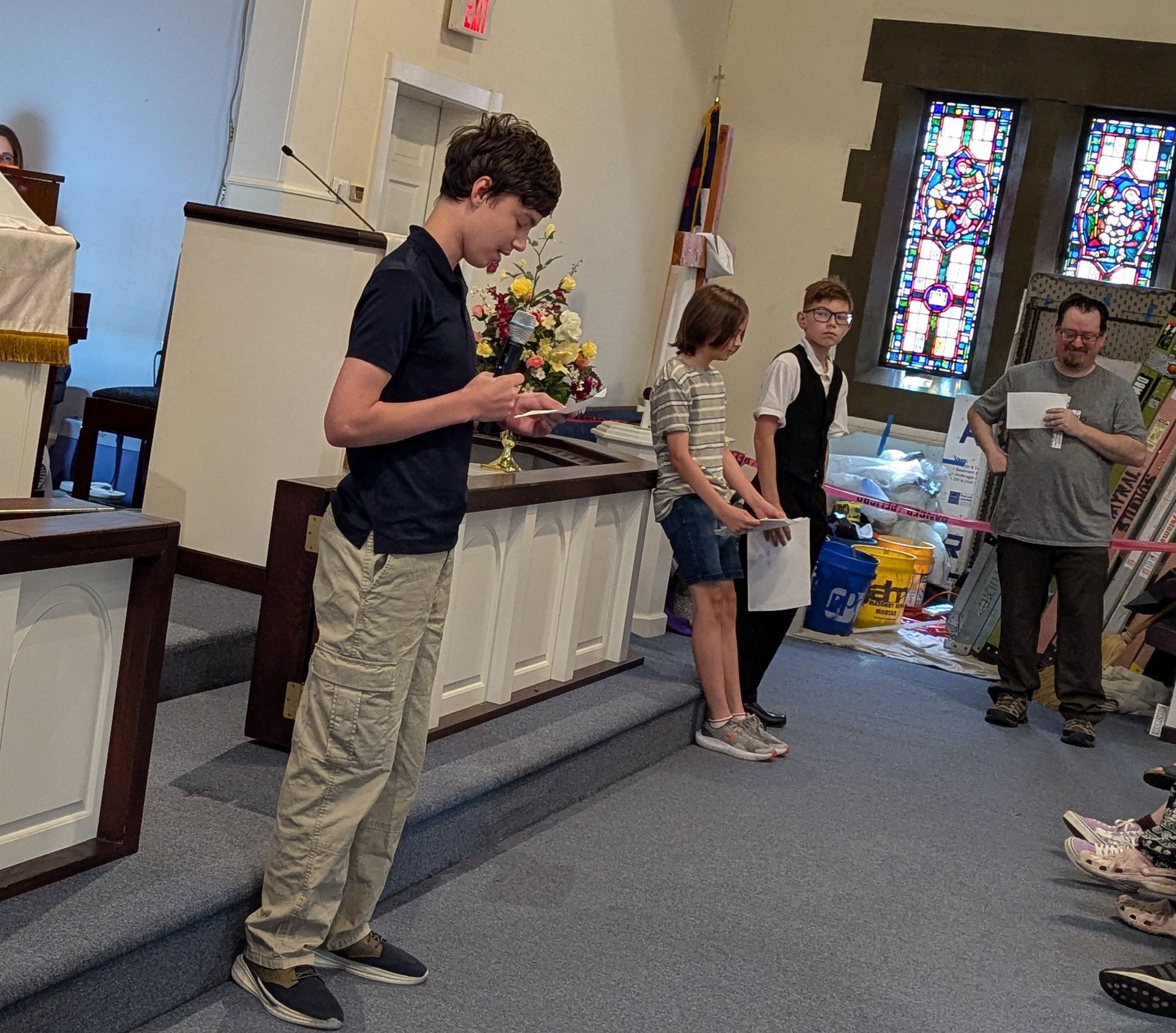 Young people on a church stage; one reads from a booklet, others stand. Sunlight streams through stained glass.