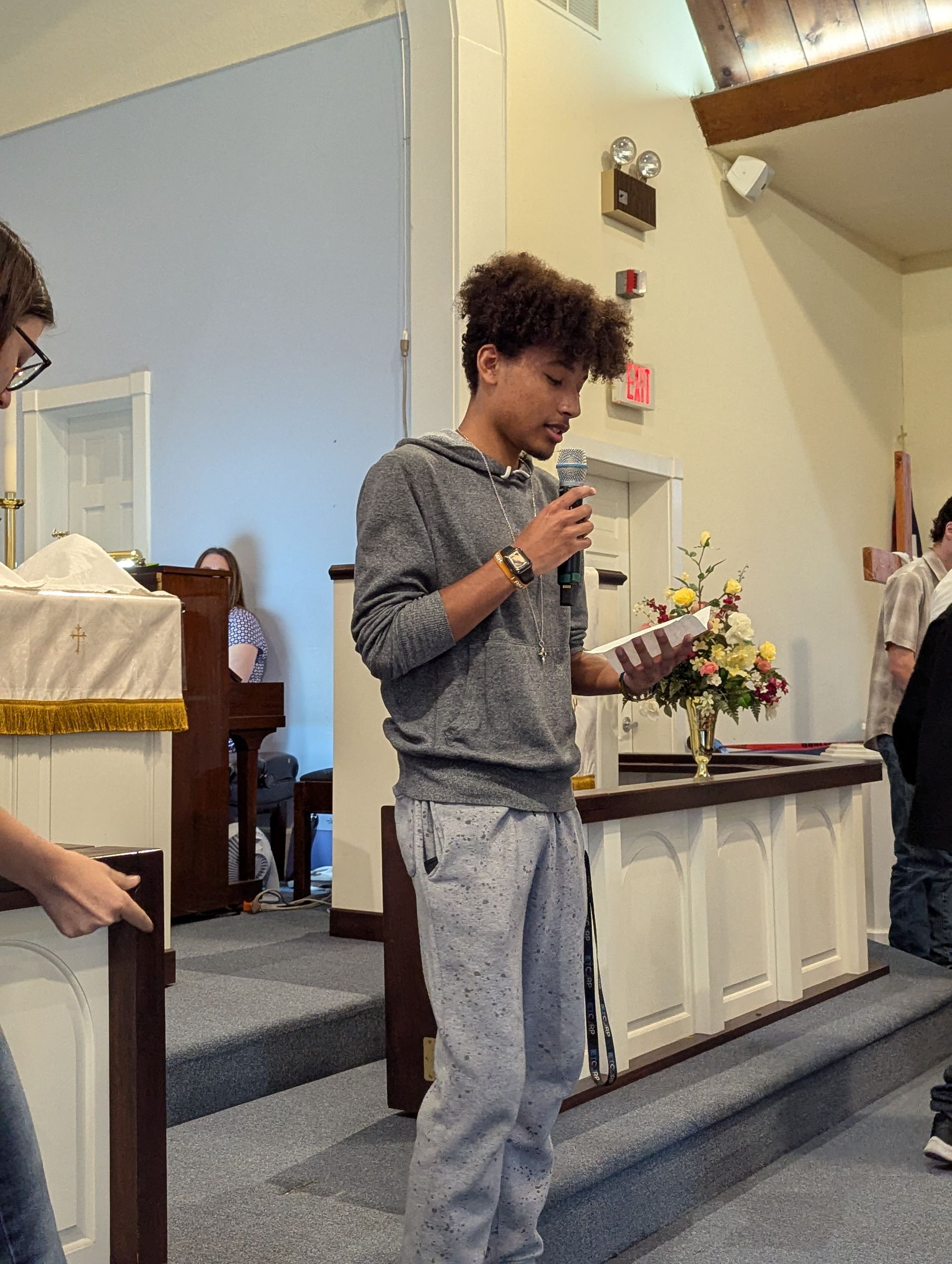 Teenager speaking into a microphone at a church, holding a paper. Flowers and a podium are visible.