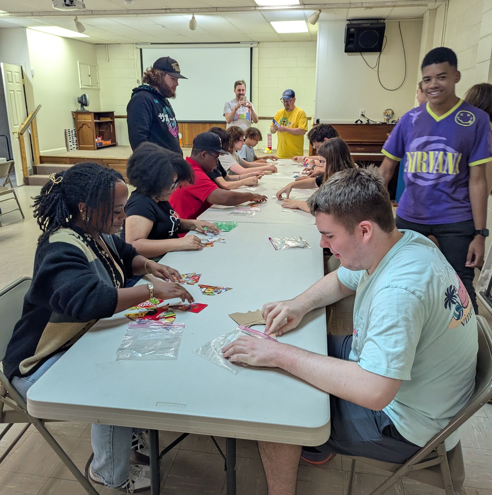 Youth at a table arraning puzzle pieces made up of cereal boxes. Adults watching. Indoor setting.