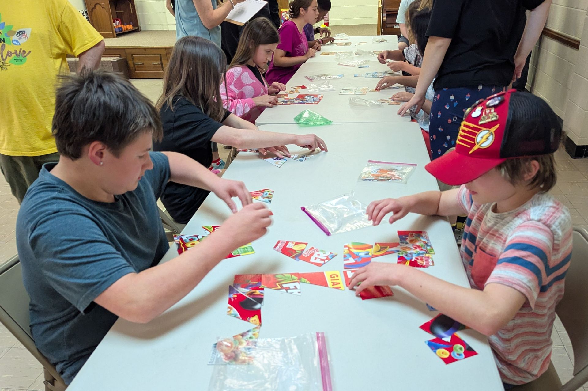 Youth arrange puzzle pieces at a long table. One boy wears a red cap. Others reach for game pieces.