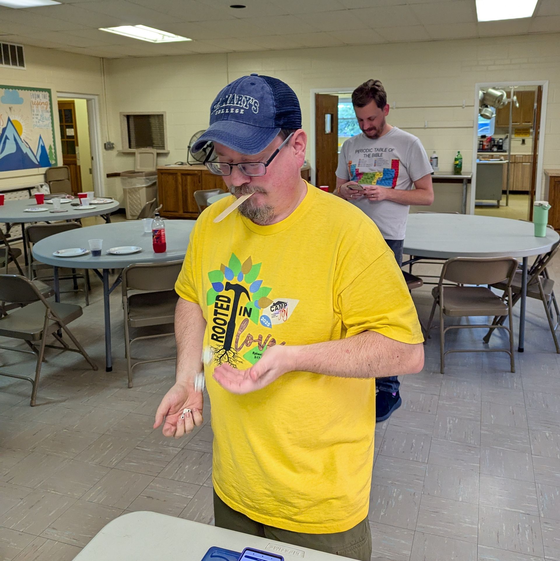 Ryan, in a yellow shirt and a blue hat, holding items, indoors with tables, and Pastor Matthew staning behind him.