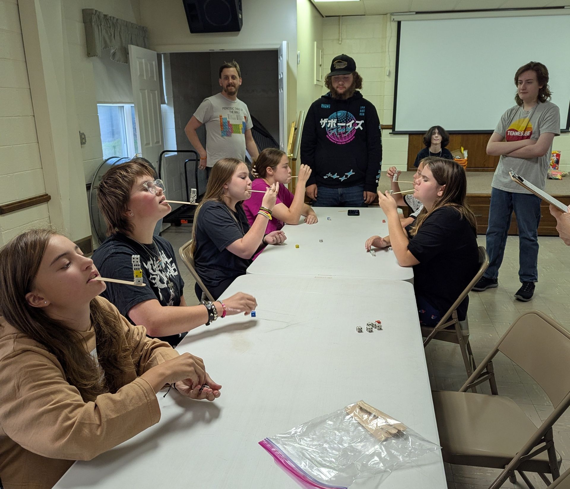 Youth playing a party game, trying to move items with straws. Indoors, a long table with participants and spectators.