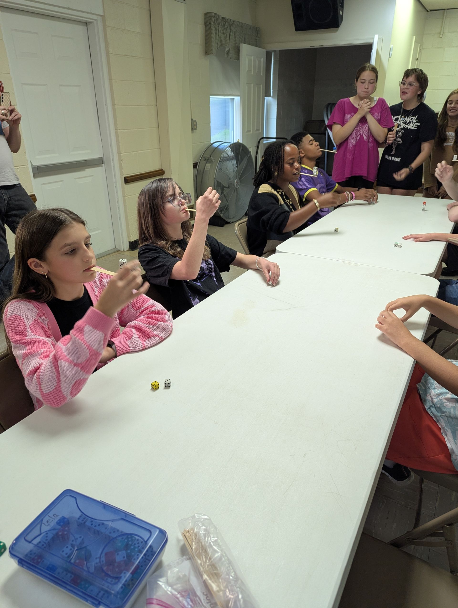 Youth playing a game at a table, some rolling dice and others watching expectantly. Indoors, neutral colors.