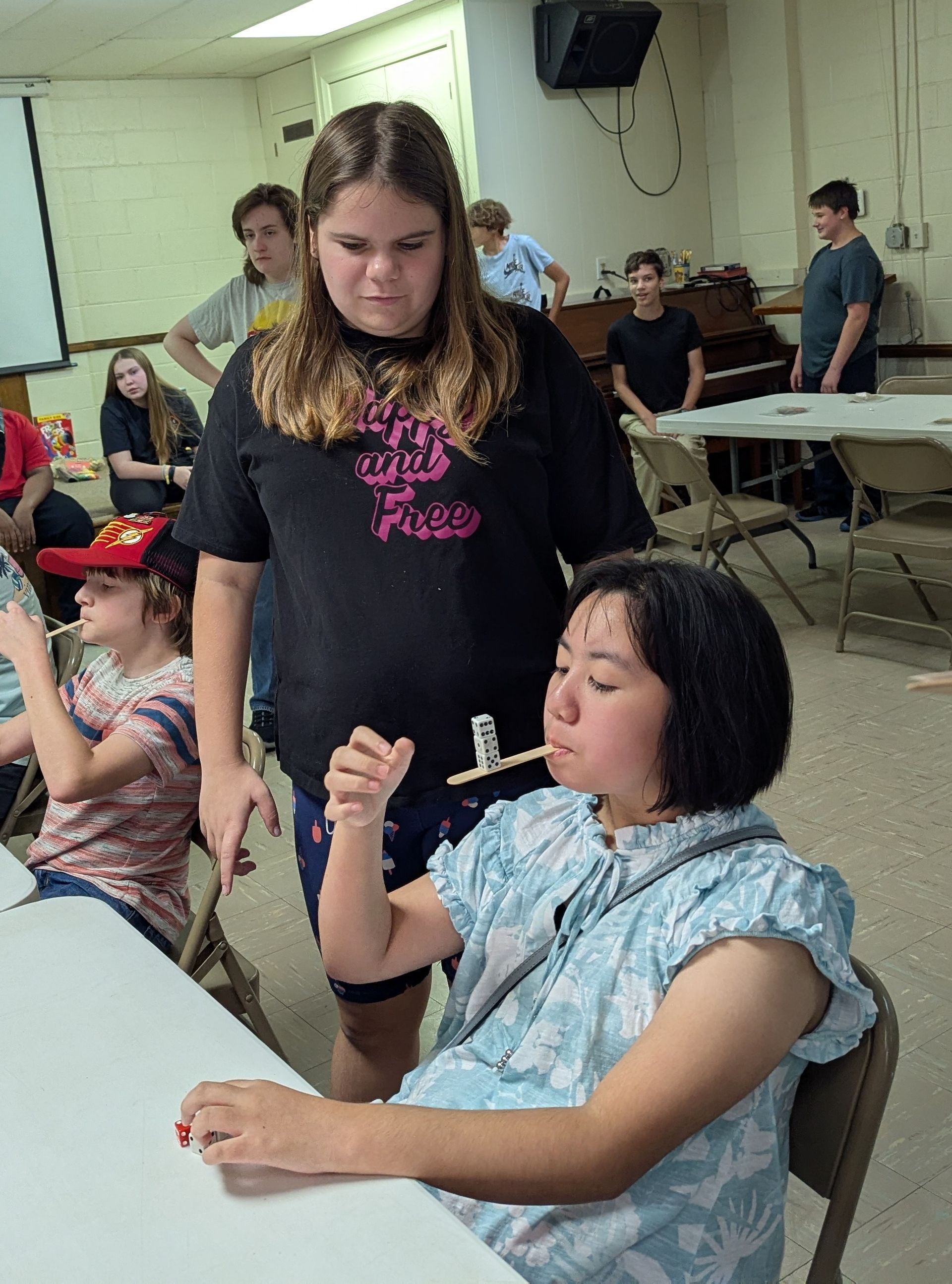 A youth balances a toothpick on her mouth while another watches. Others look on in a room.