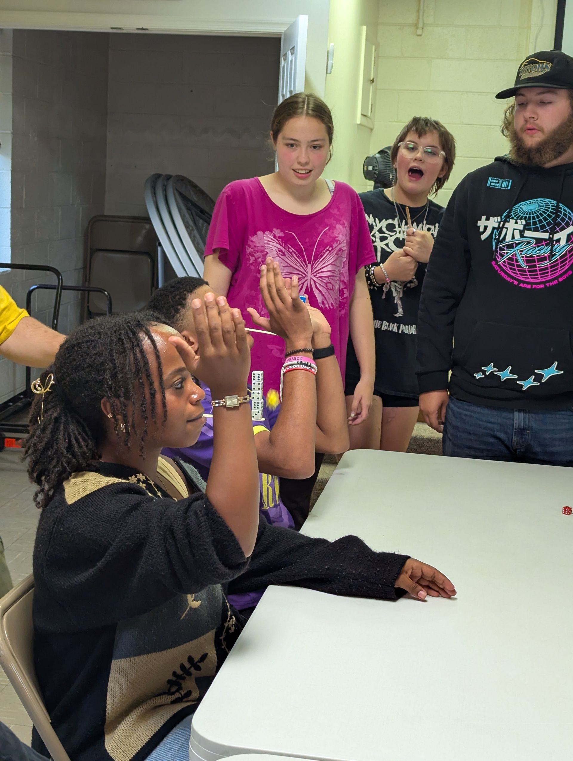Group of people gathered around a table. One girl claps, others watch with excited expressions in a room.