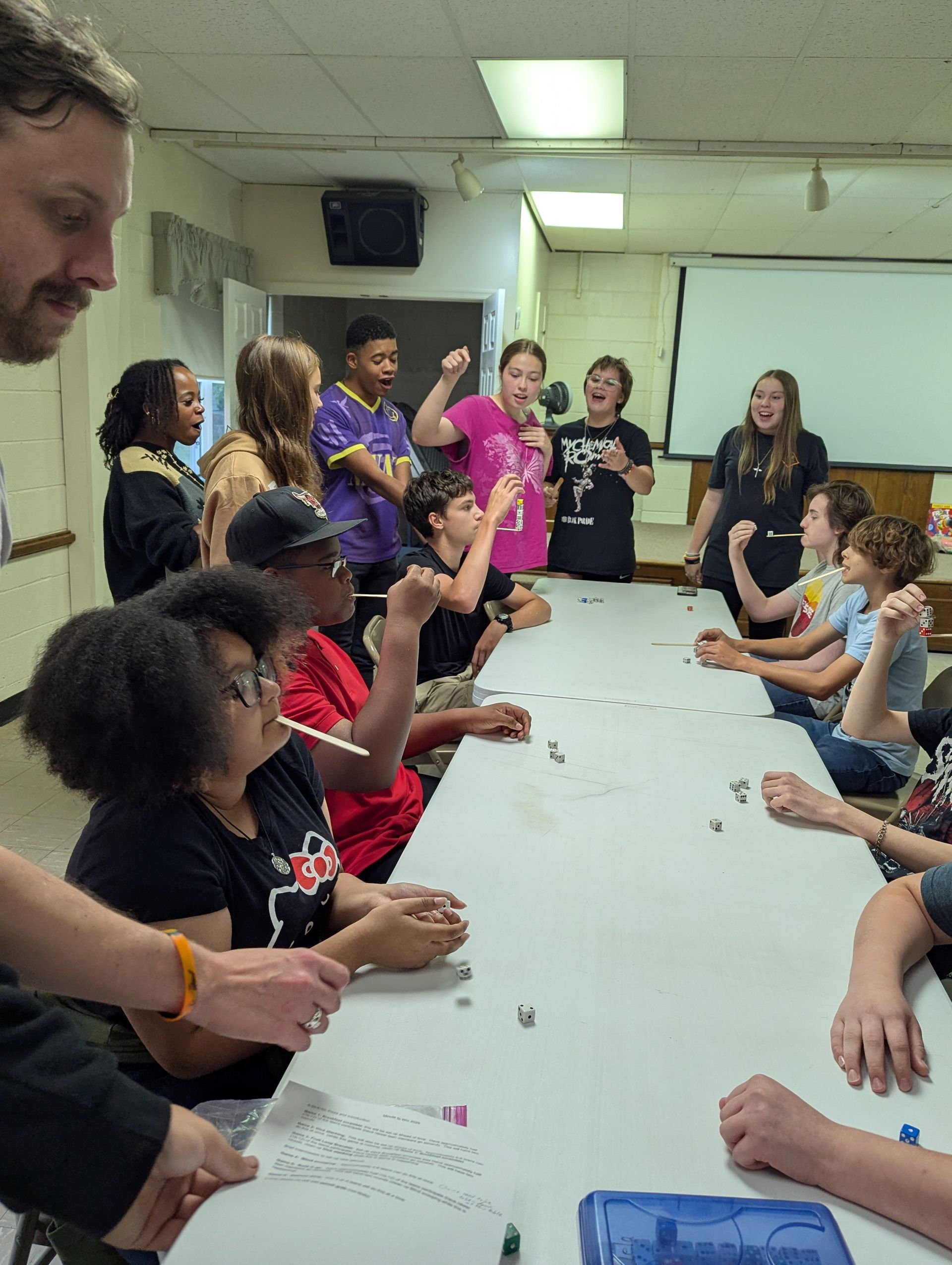 Youth and adults at a long table; some hold objects, others cheer; indoor setting.
