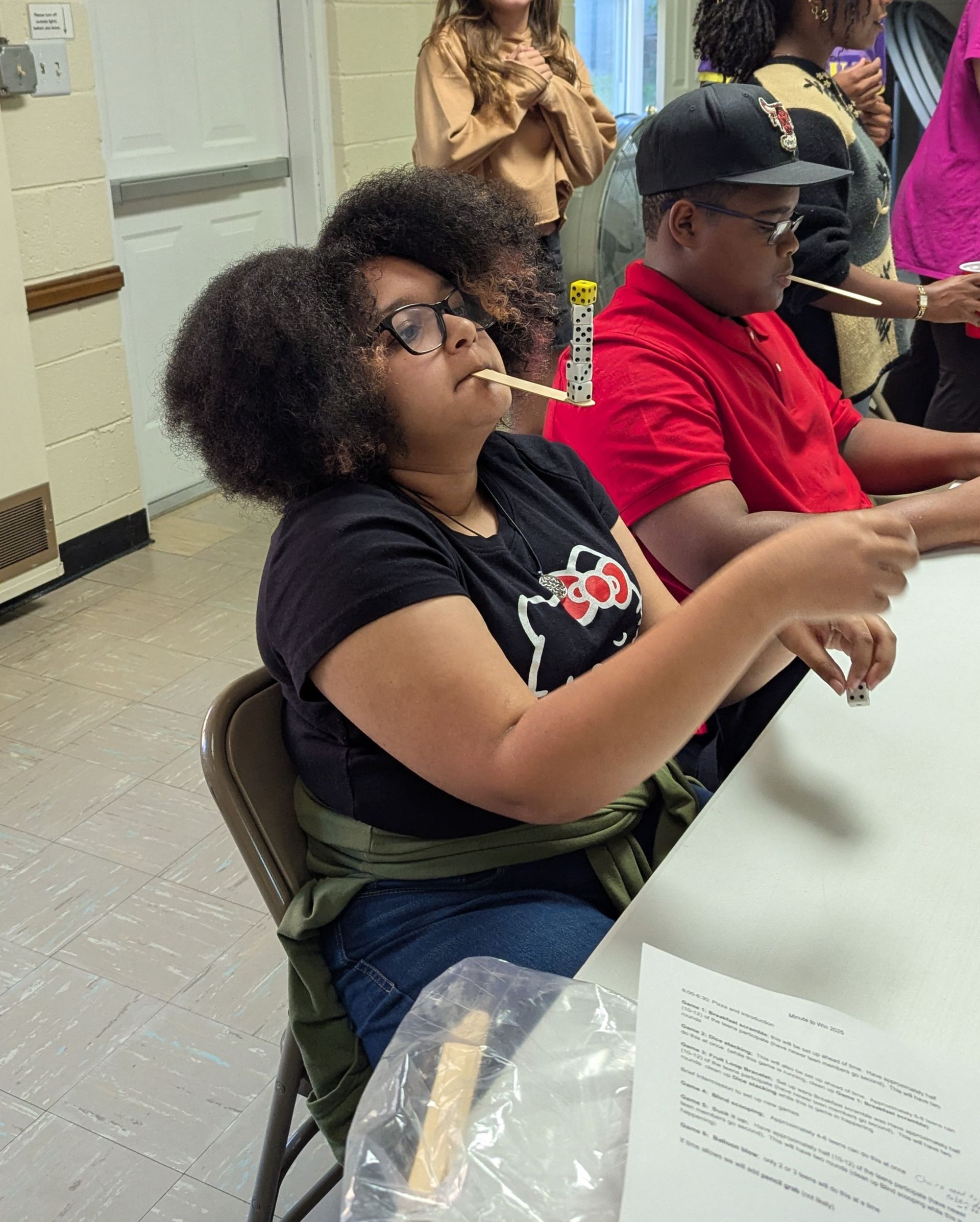 A youth balances a stick on her upper lip. Others watch around a table.