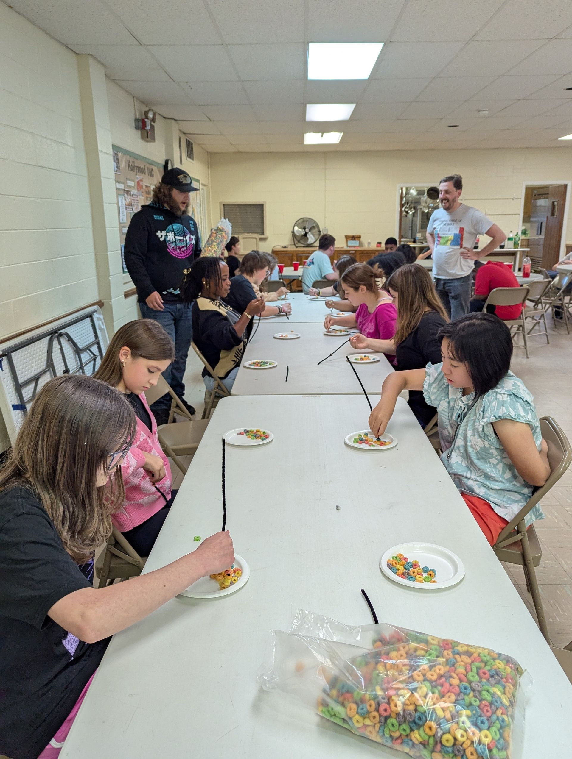 Youth painting at a long table. Pastor Matthew supervises. Colorful cereal in a bag. Indoor setting.