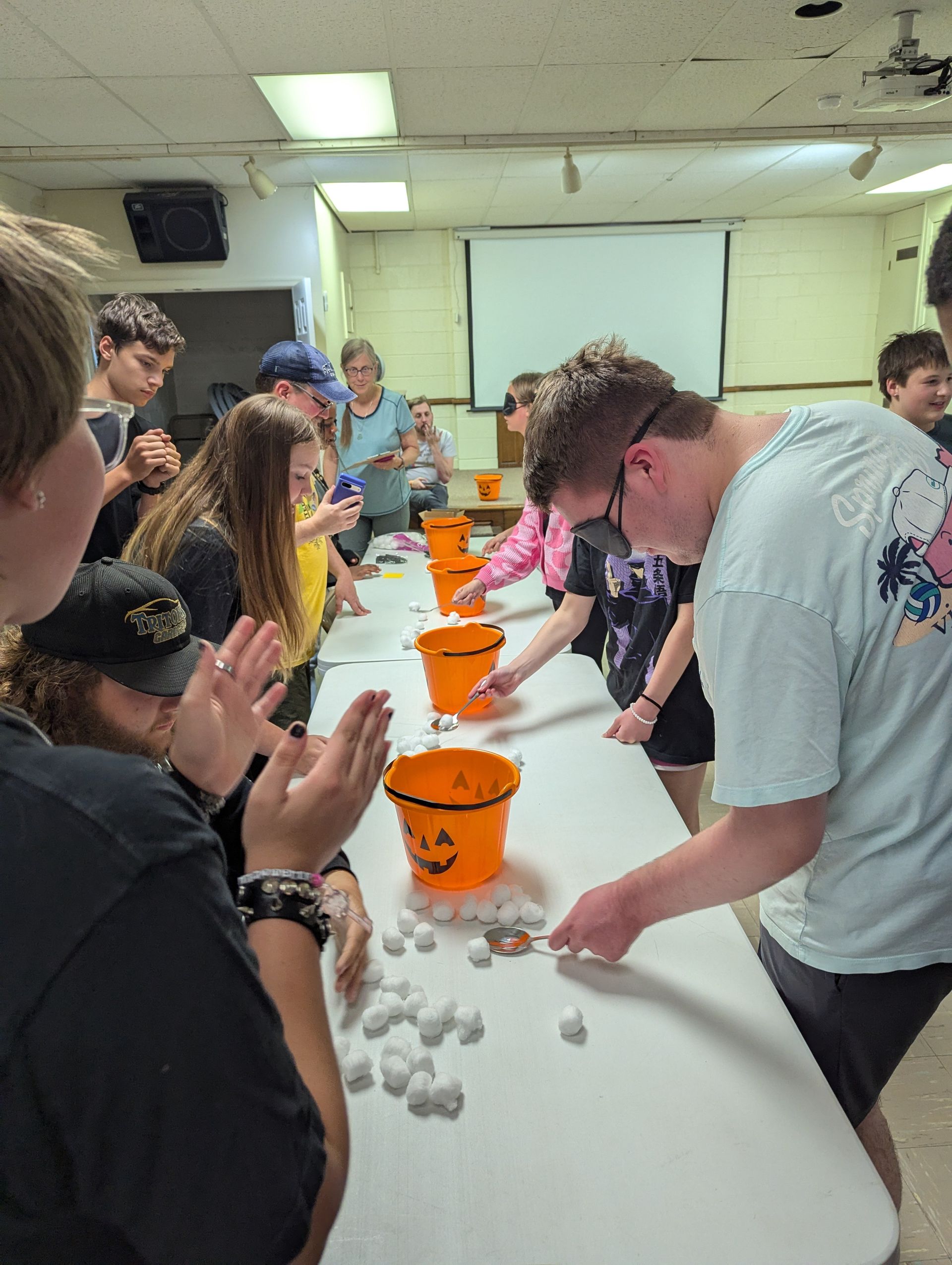 Youth playing a game, trying to throw items into orange Halloween buckets on a long table indoors.