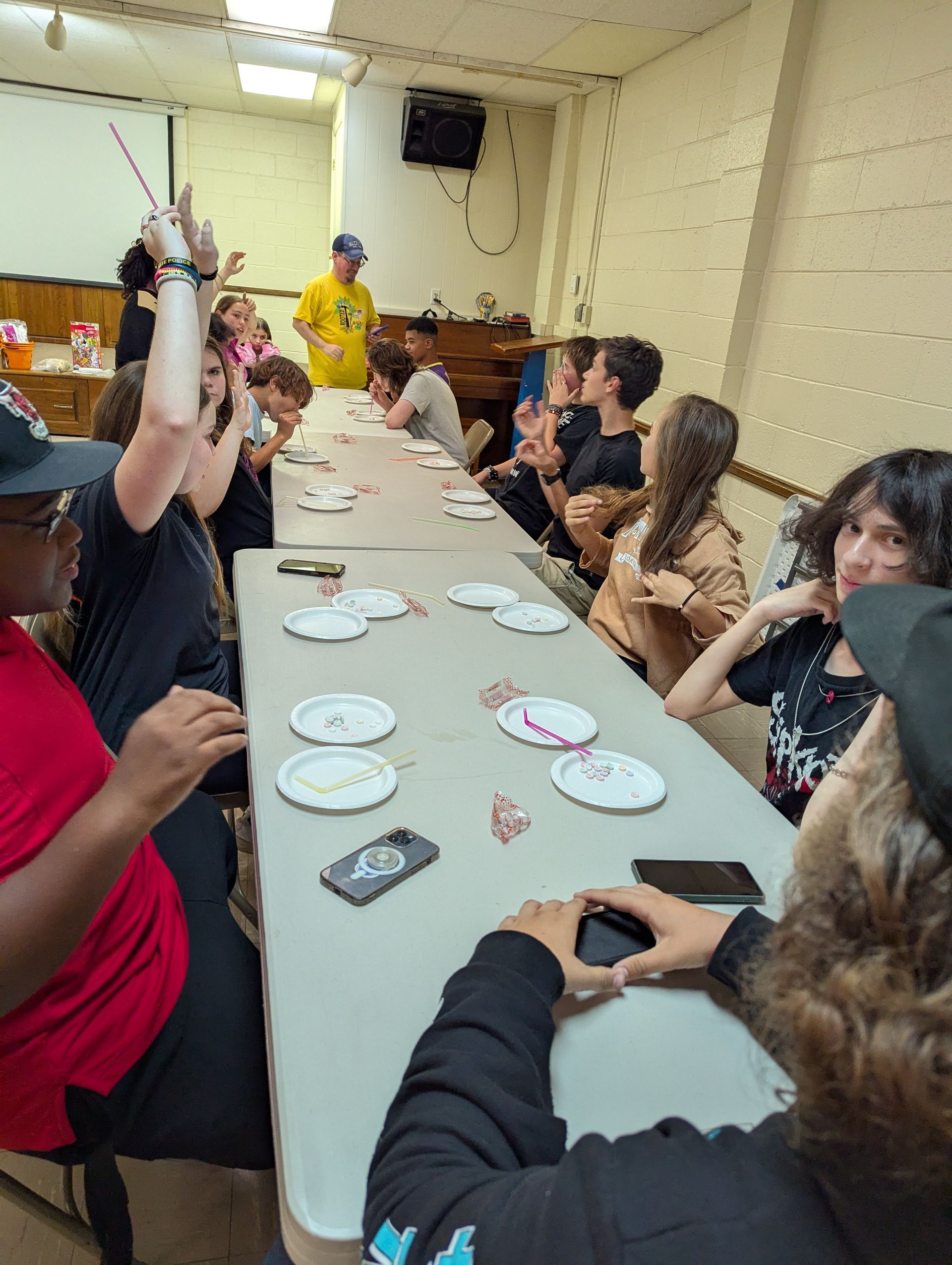 A group of teens at a long table, some raising hands excitedly, playing a game.