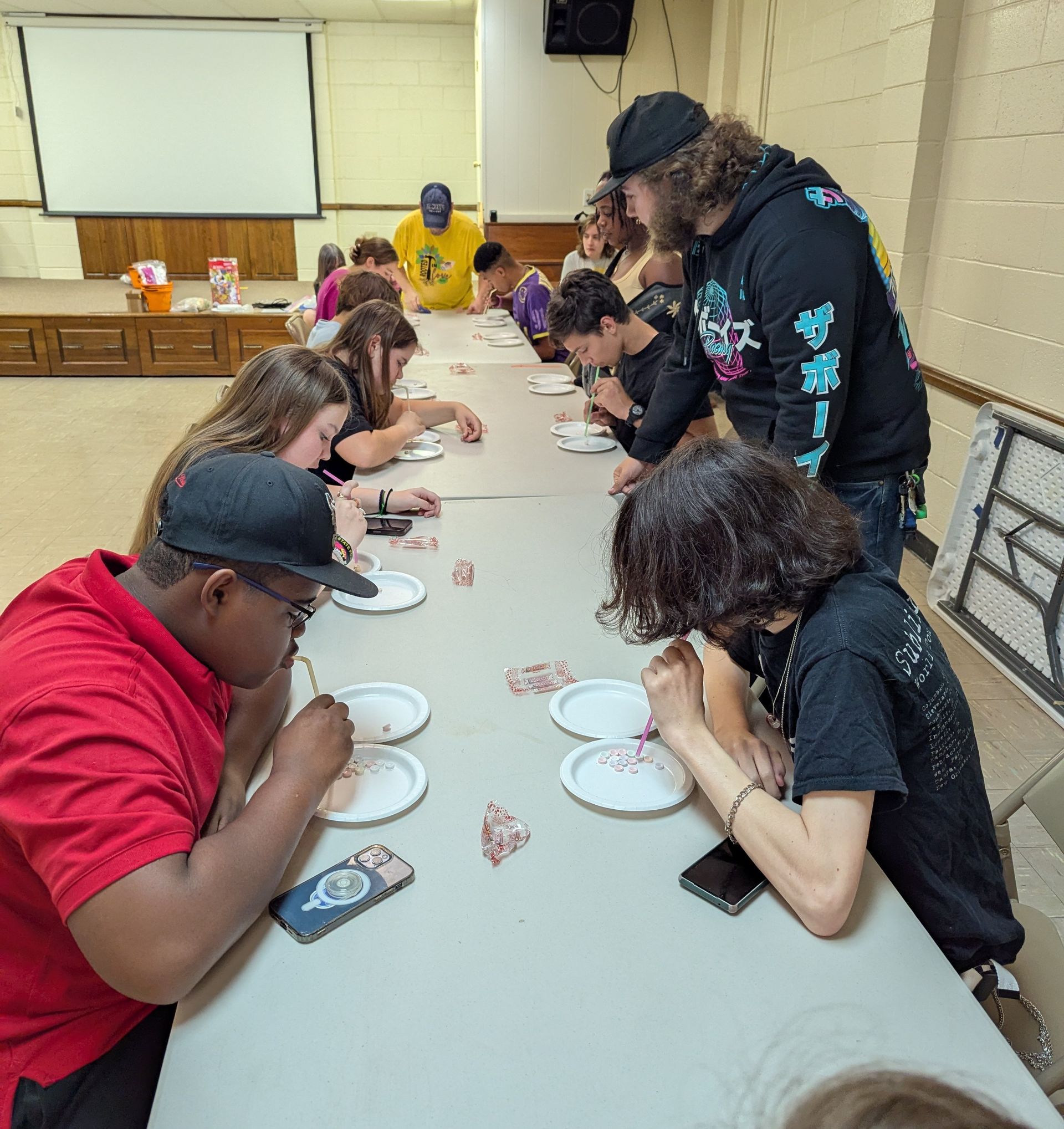 A group of teens at a long table, competing in a game, some eating from small plates.