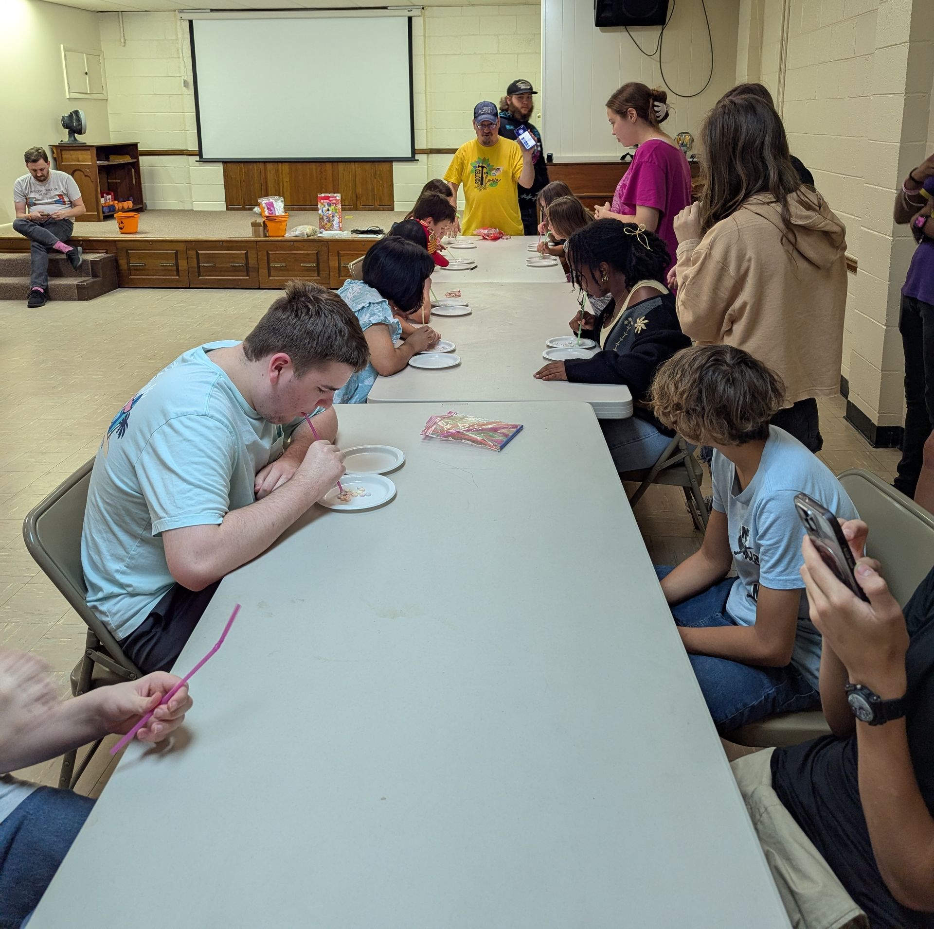 Youth at a table, competing. Some use straws, snacks on plates. Indoors, with a projector screen.