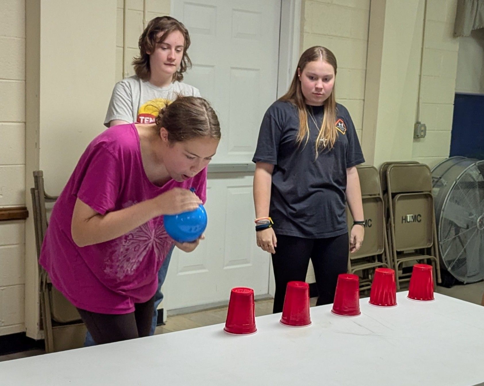 Three people playing a game. A girl blows a blue ball towards red cups on a table. Two other girls watch.