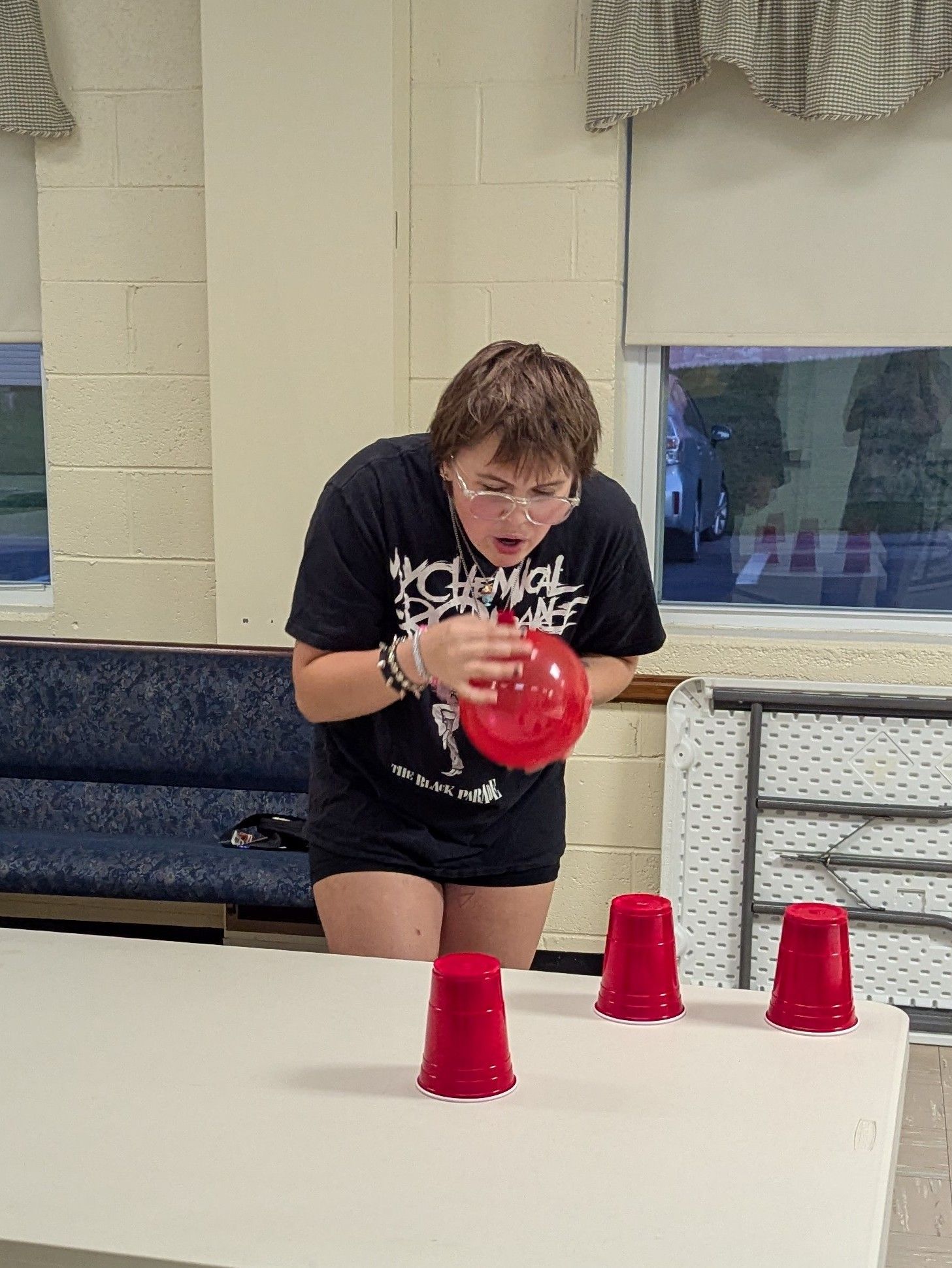 Youth blowing a red balloon toward red cups on a table. Indoors, looking focused, playing a game.