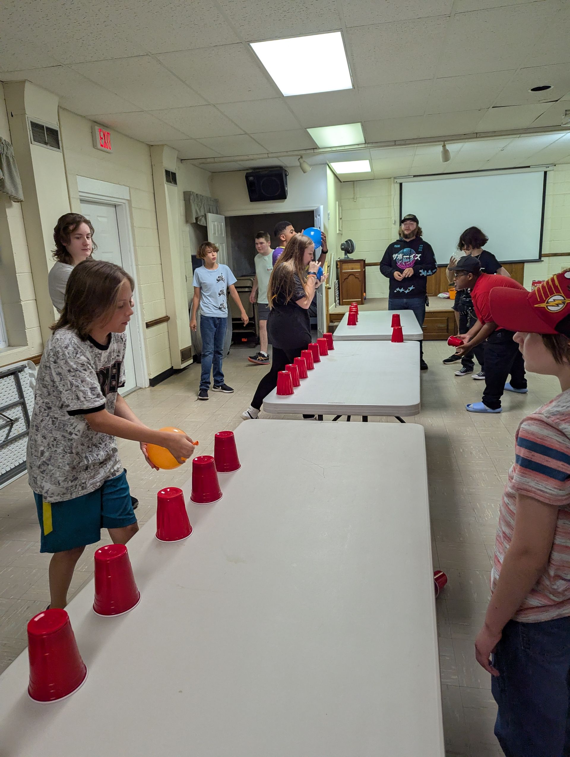 Youth playing a game with red cups and a ball. Indoors, with tables, a projector screen, and other kids watching.