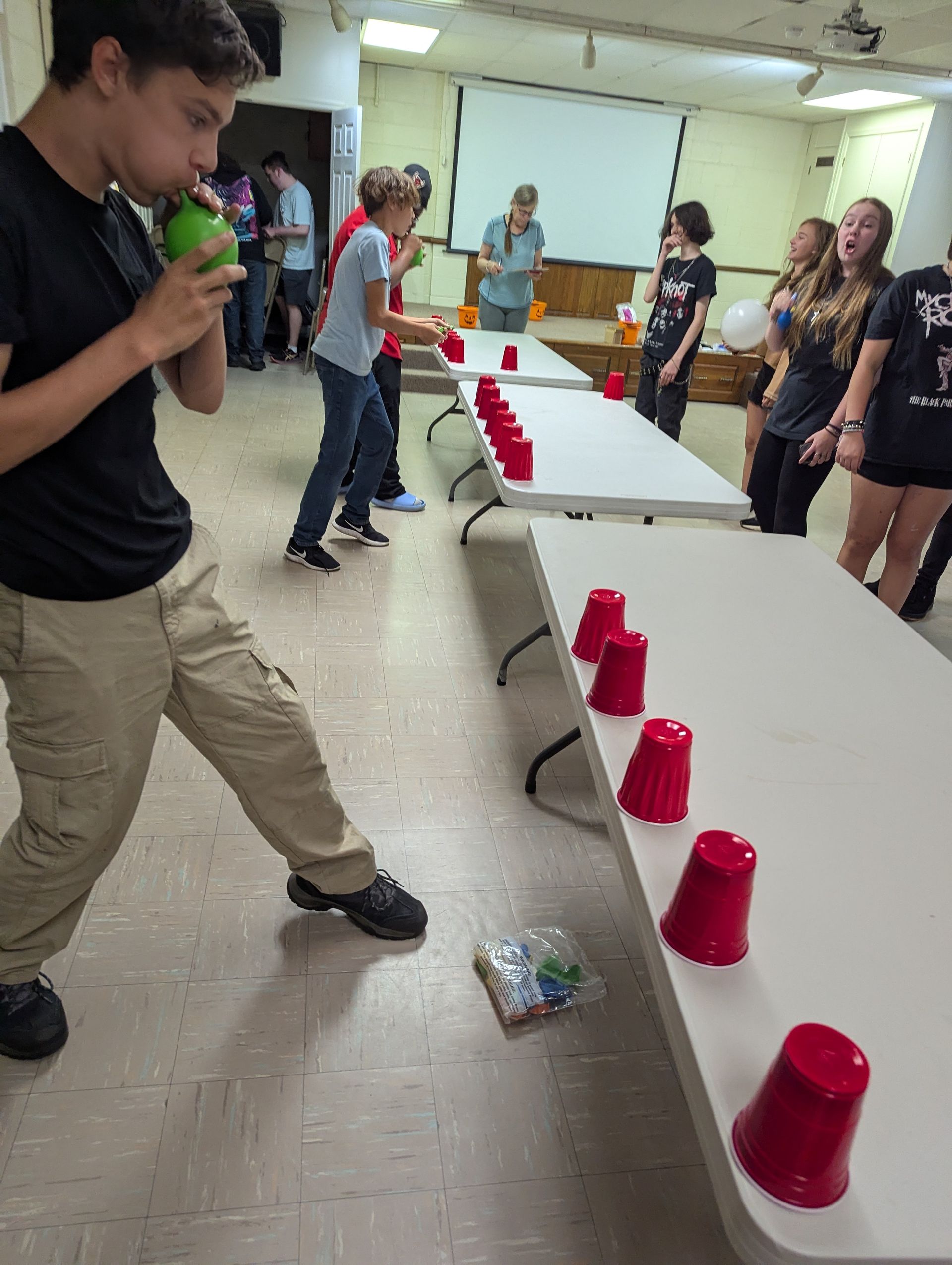Group playing a game; teens blowing ping pong balls toward red cups on tables.