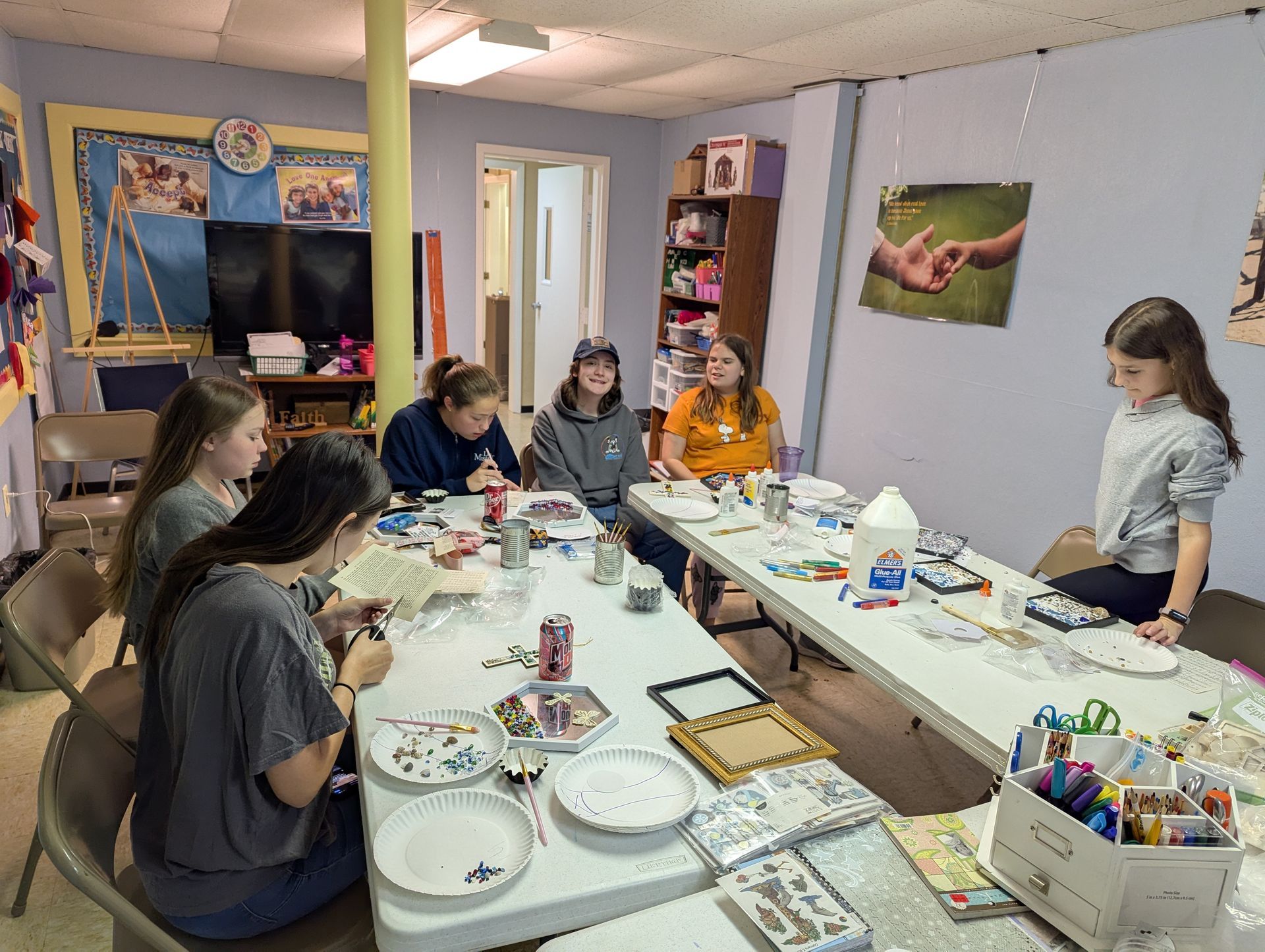 A group of teens craft around tables, with art supplies in a brightly lit room.