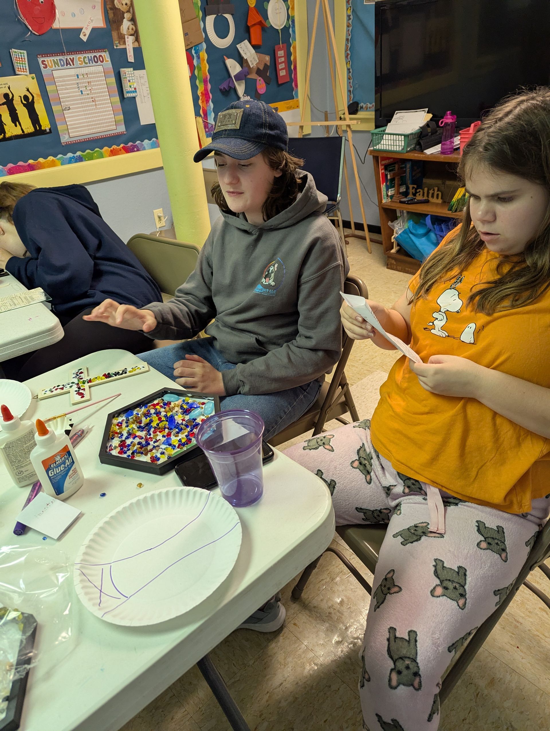 Two youth craft at a table: one in grey hoodie and blue cap, the other in orange shirt, wearing pajamas.