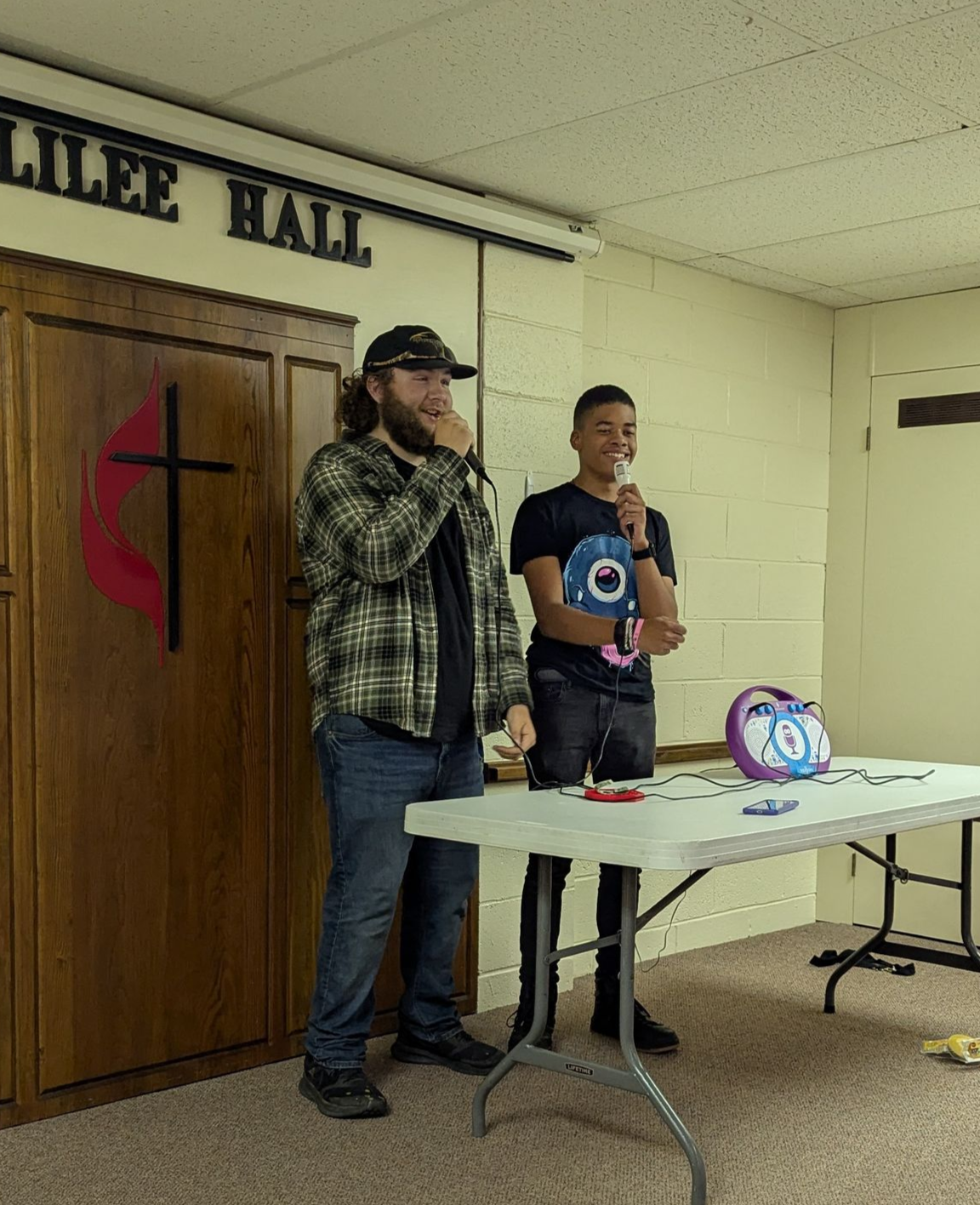 Two young men singing at a table in front of a door labeled