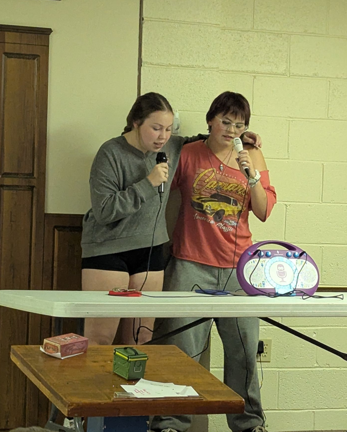 Two young women singing karaoke into microphones at a table, with a stereo.
