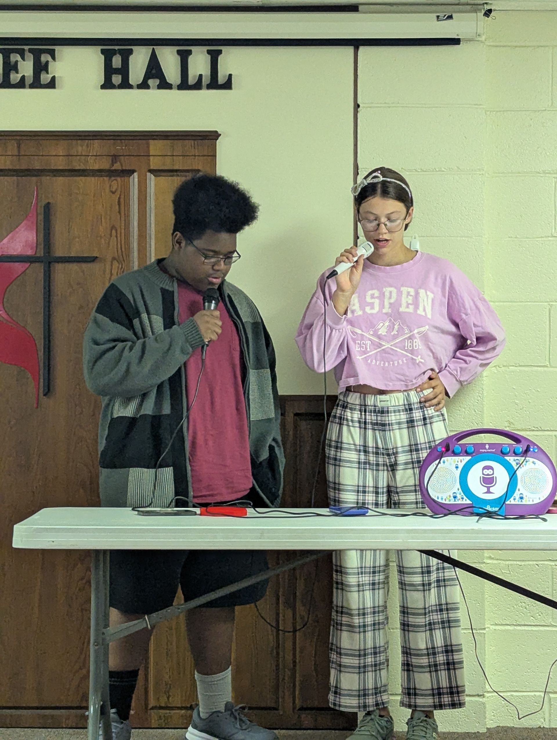 Two teens sing into mics at a table in a hall. One wears a sweater, shorts. The other a long-sleeved shirt, plaid pants.
