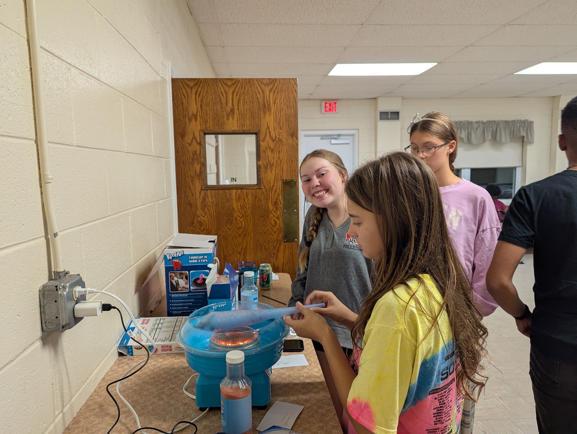 Youth making a cotton candy cone. Two girls smiling, looking at the machine. Indoors, lit by overhead lights.