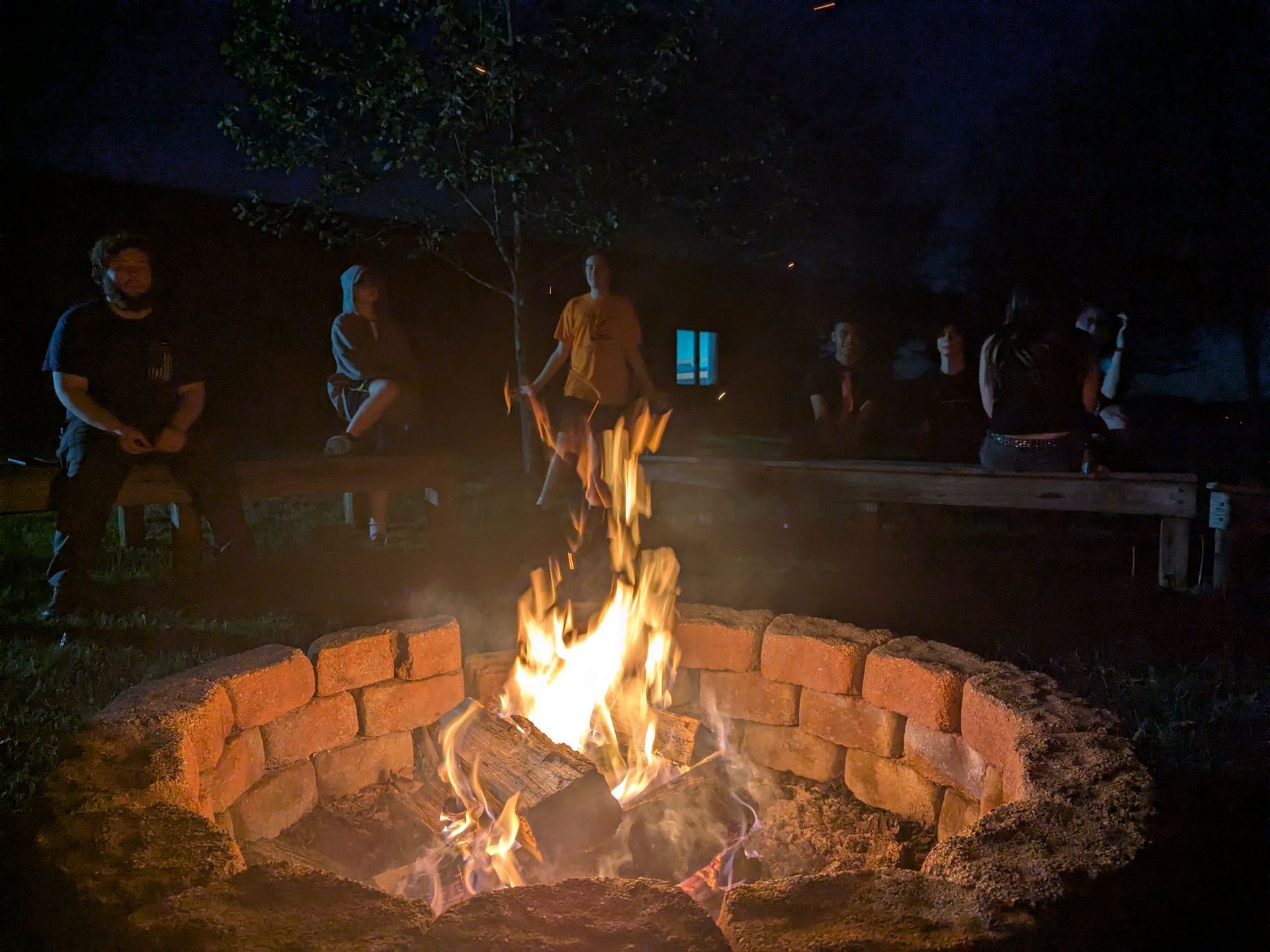 People gathered around a brick fire pit at night, with flames lighting the dark outdoor setting.